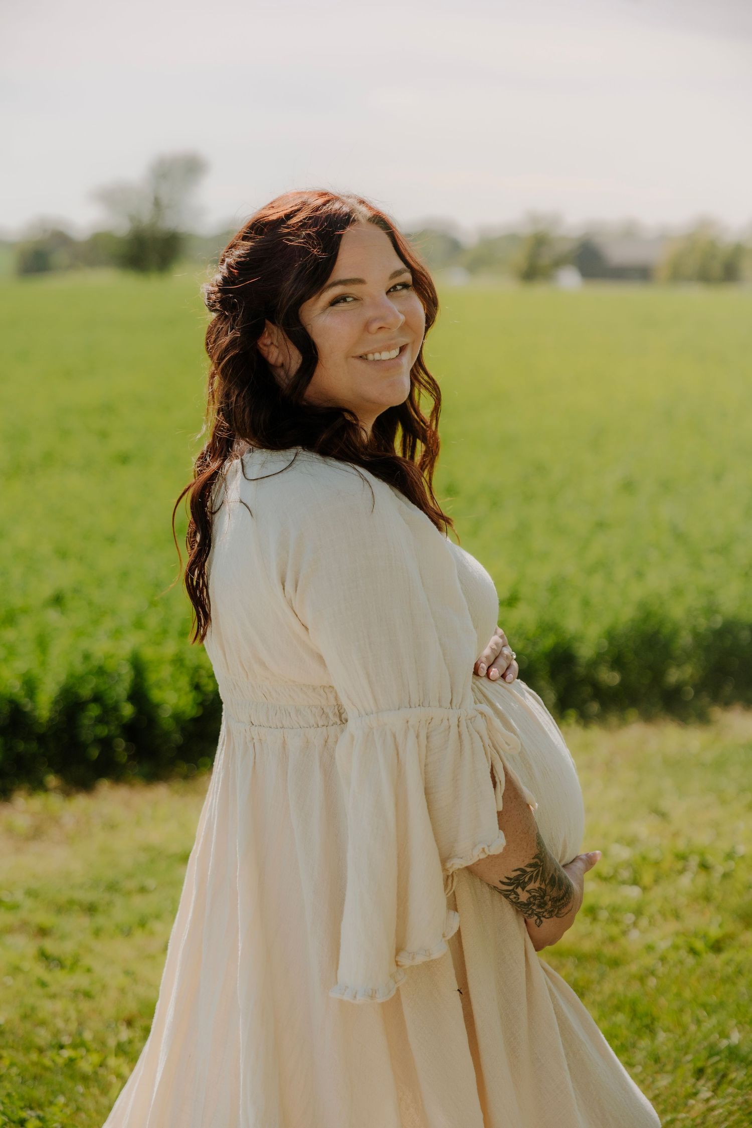 Smiling pregnant woman in cream dress holding belly in green field, maternity photography outdoors.