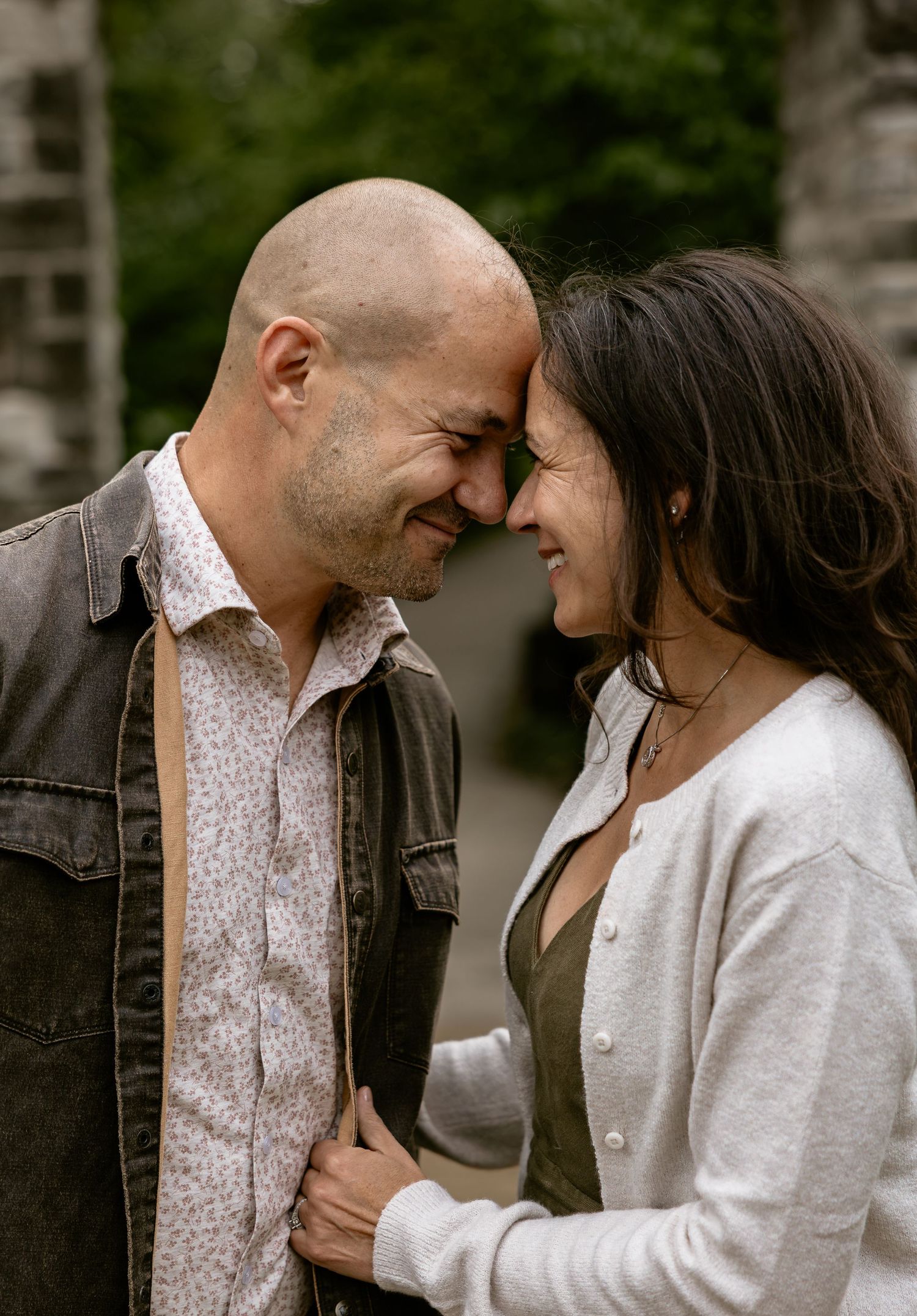 A bald man and brunette woman smile and touch foreheads in an affectionate outdoor couples portrait.
