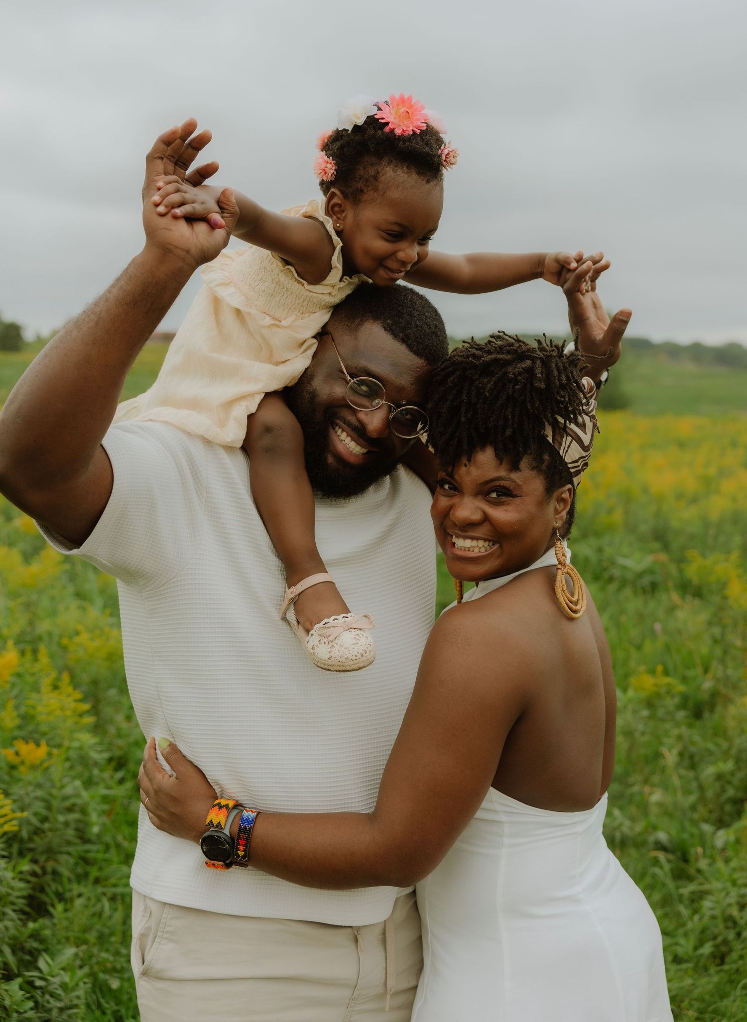 Happy Black family posing outdoors in a green field, father holding toddler on shoulders while mother embraces them, smiling.