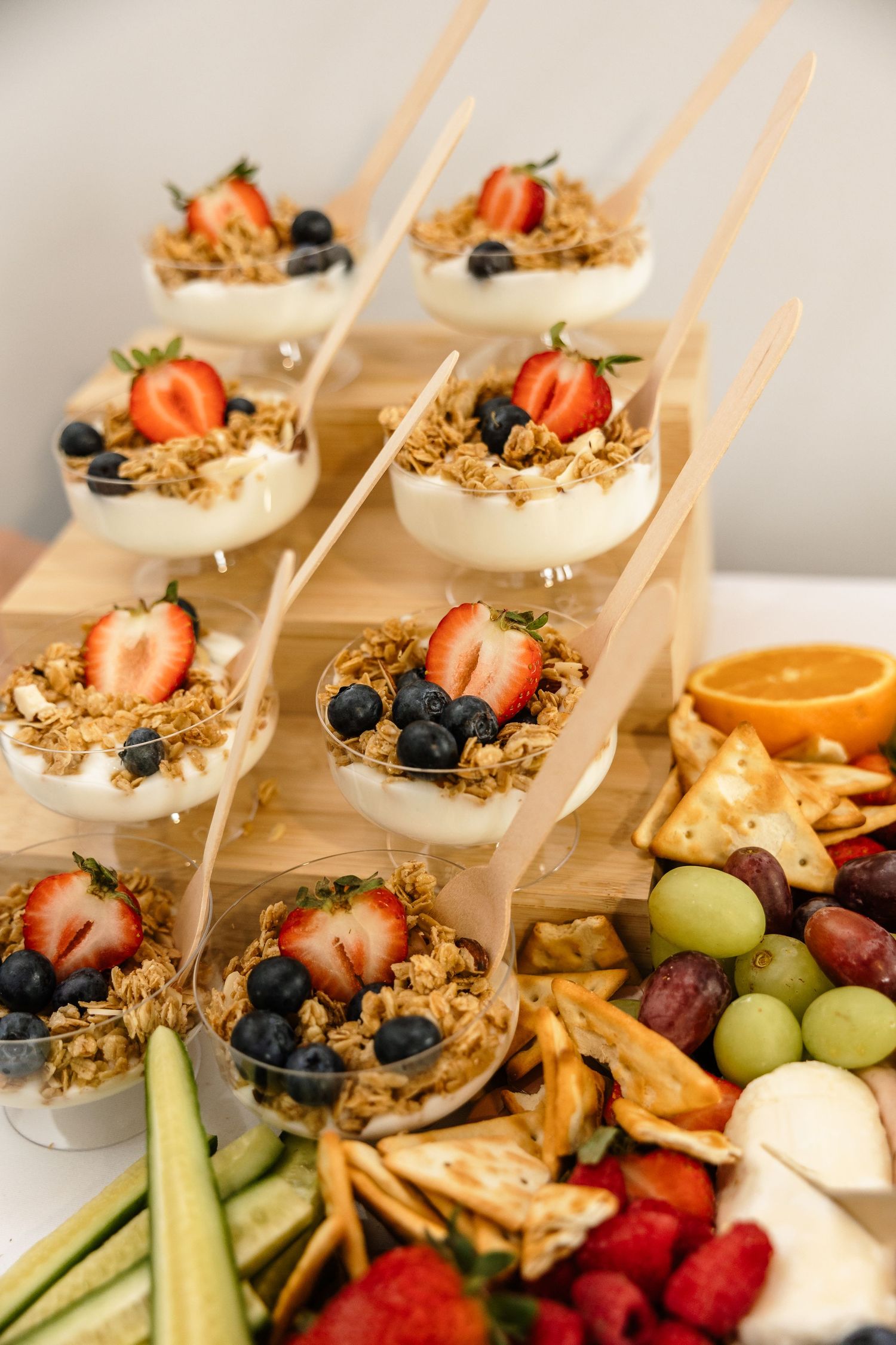 Elegant yogurt parfait cups with granola, strawberries, and blueberries on a wooden stand beside a fresh fruit and cracker platter.