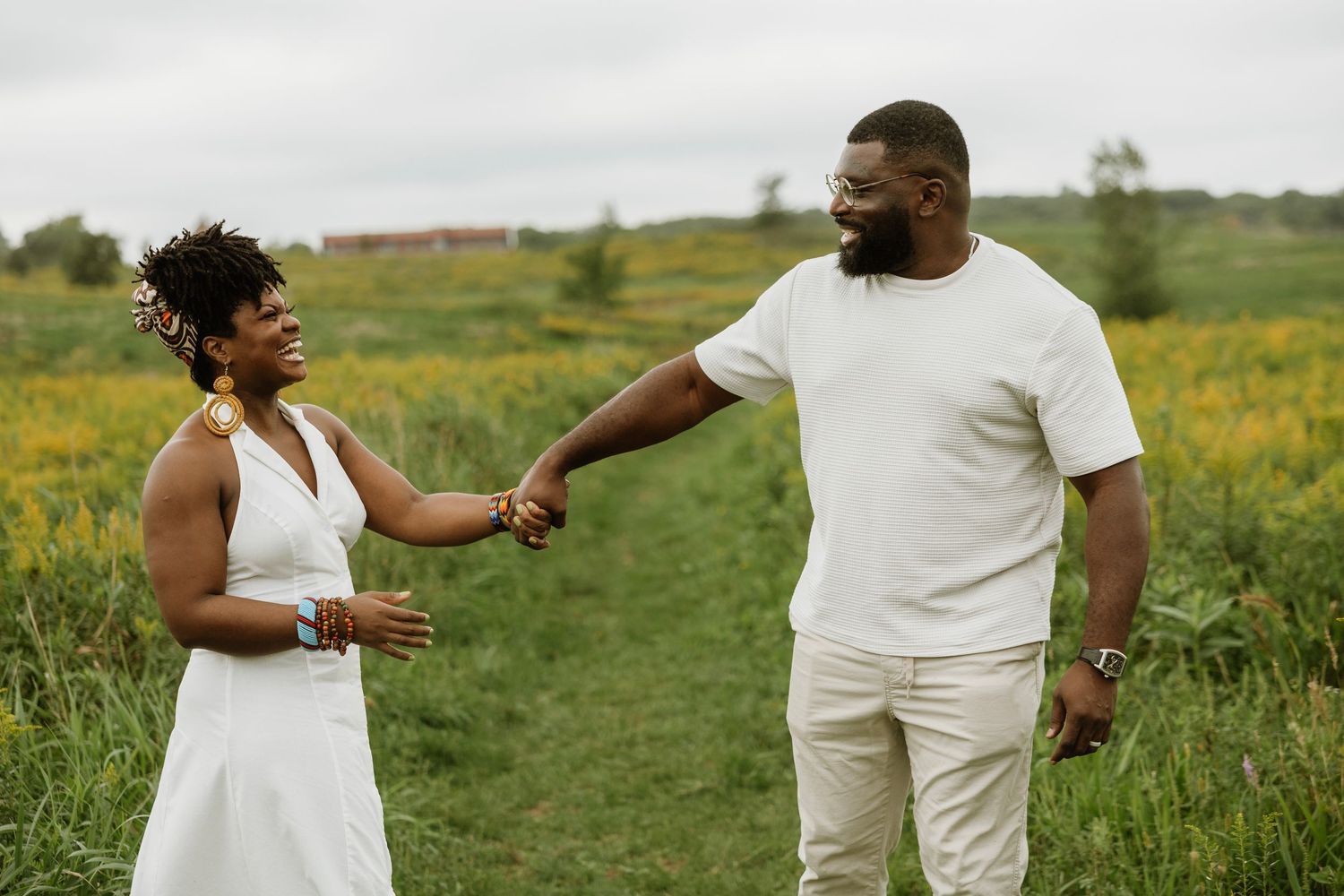 A joyful Black couple holds hands in a lush green meadow, the woman in white dress, man in white outfit.