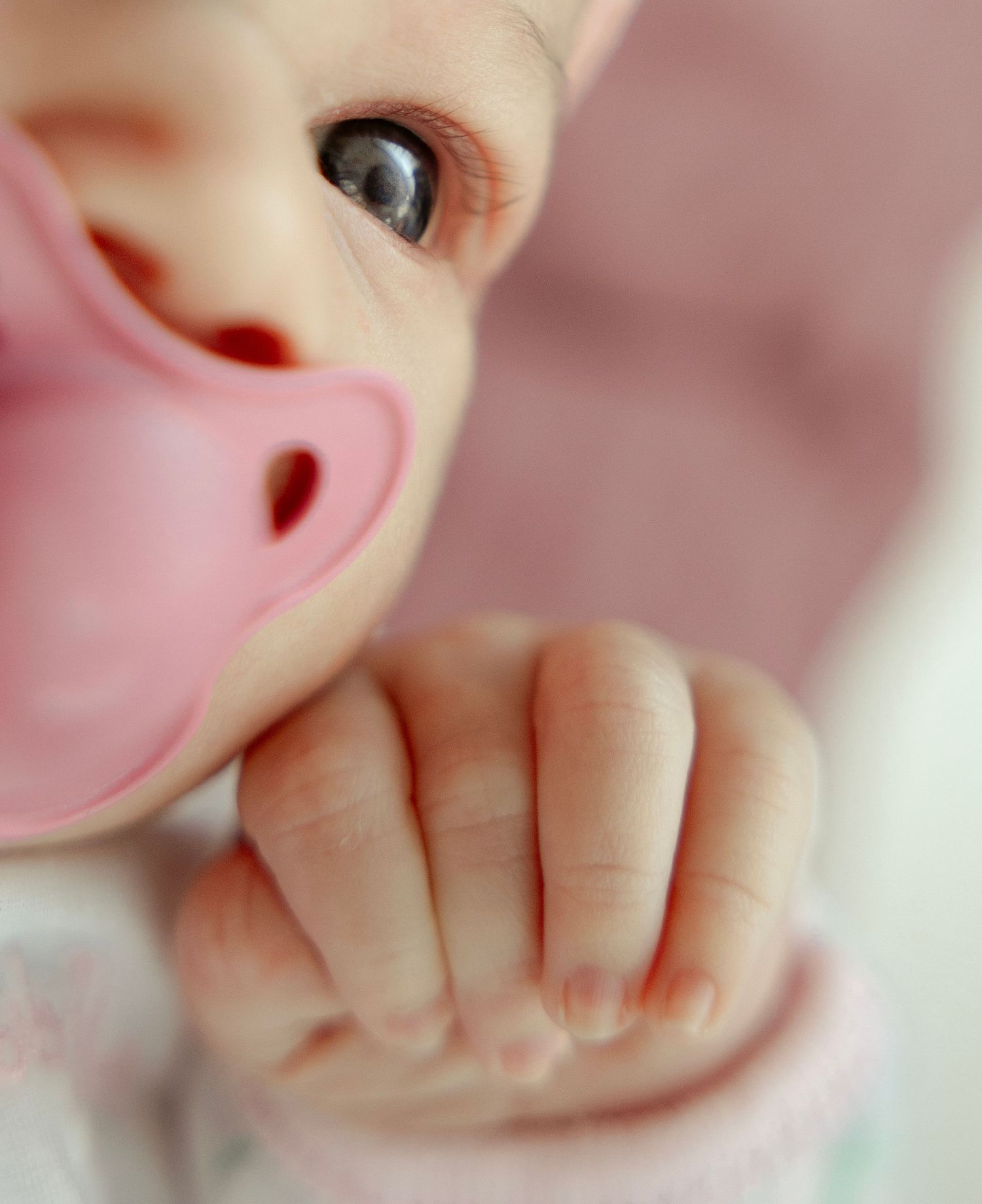 Close-up of a newborn baby with blue eyes holding a pink pacifier in their mouth.