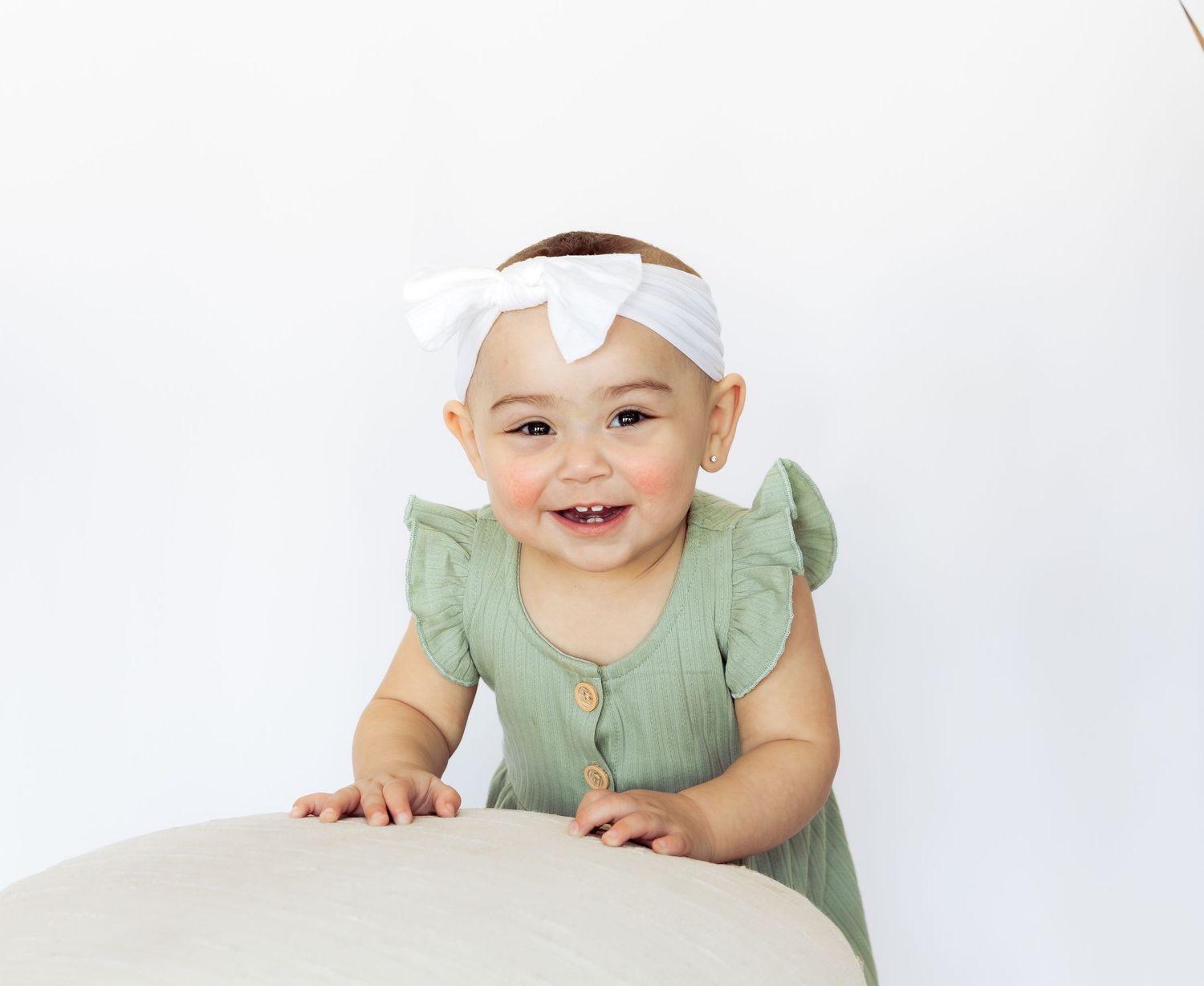Smiling baby girl in green ruffle romper and white bow headband standing at a table against white background.