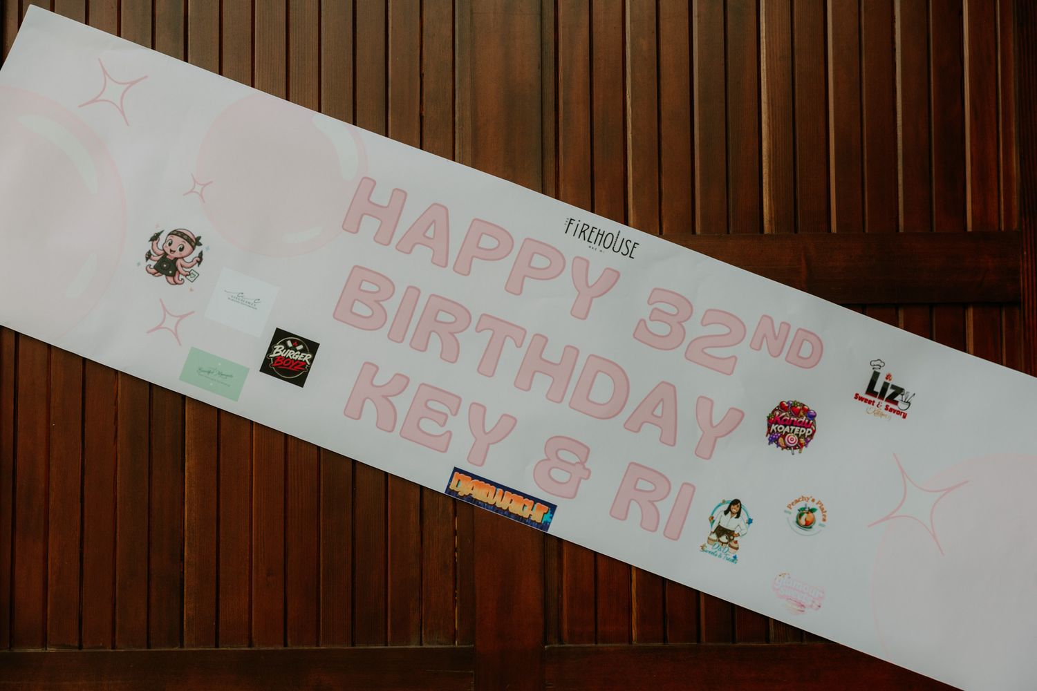 A white birthday banner reading Happy 32nd Birthday Key and Ri with colorful logos on a wooden wall.