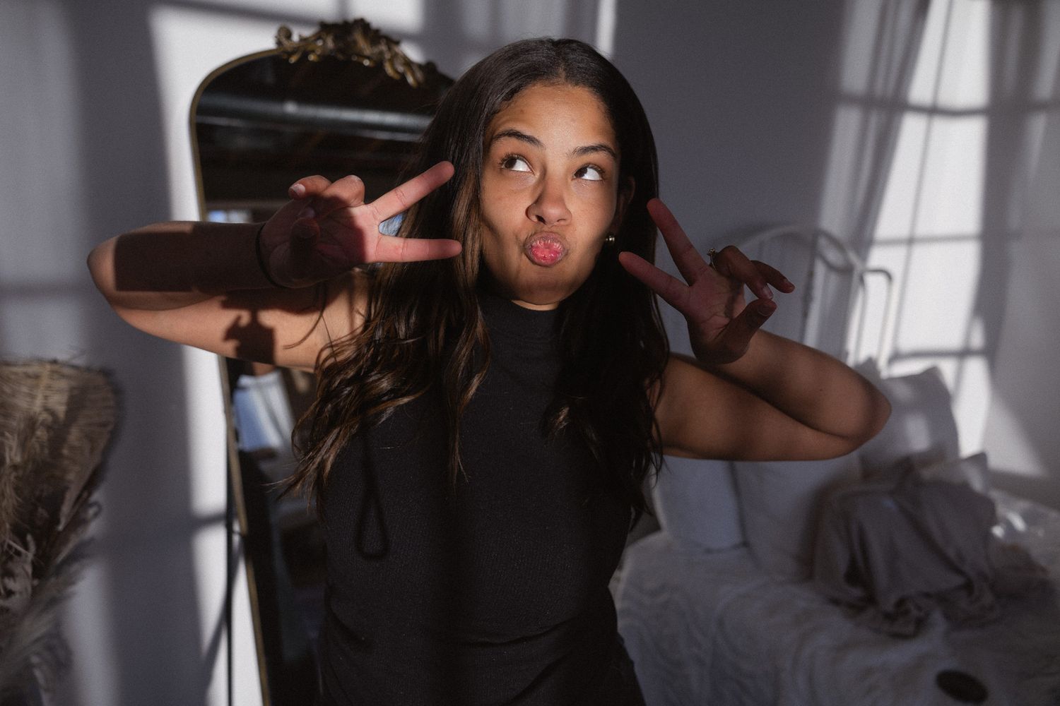 Young woman making a peace sign and duck face pose in a sunlit bedroom wearing a black sleeveless top.