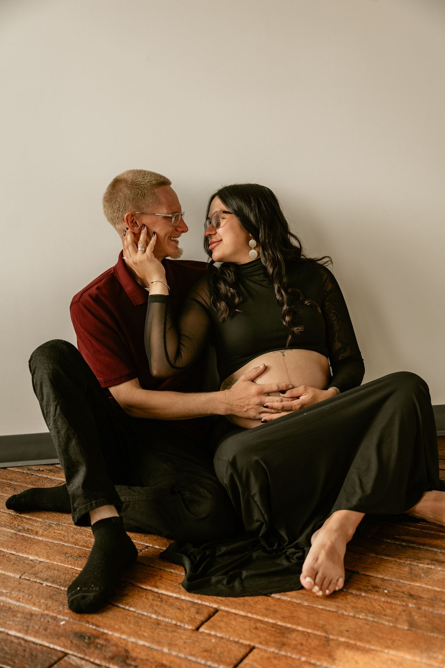 Couple sharing a tender moment during maternity photo session, pregnant woman in black dress seated on wooden floor.