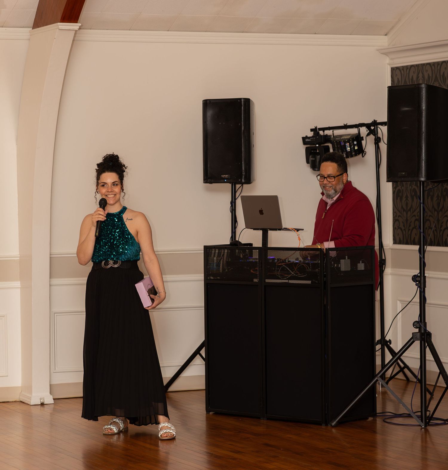 Woman in teal top speaks into microphone while DJ operates equipment at elegant indoor event venue.