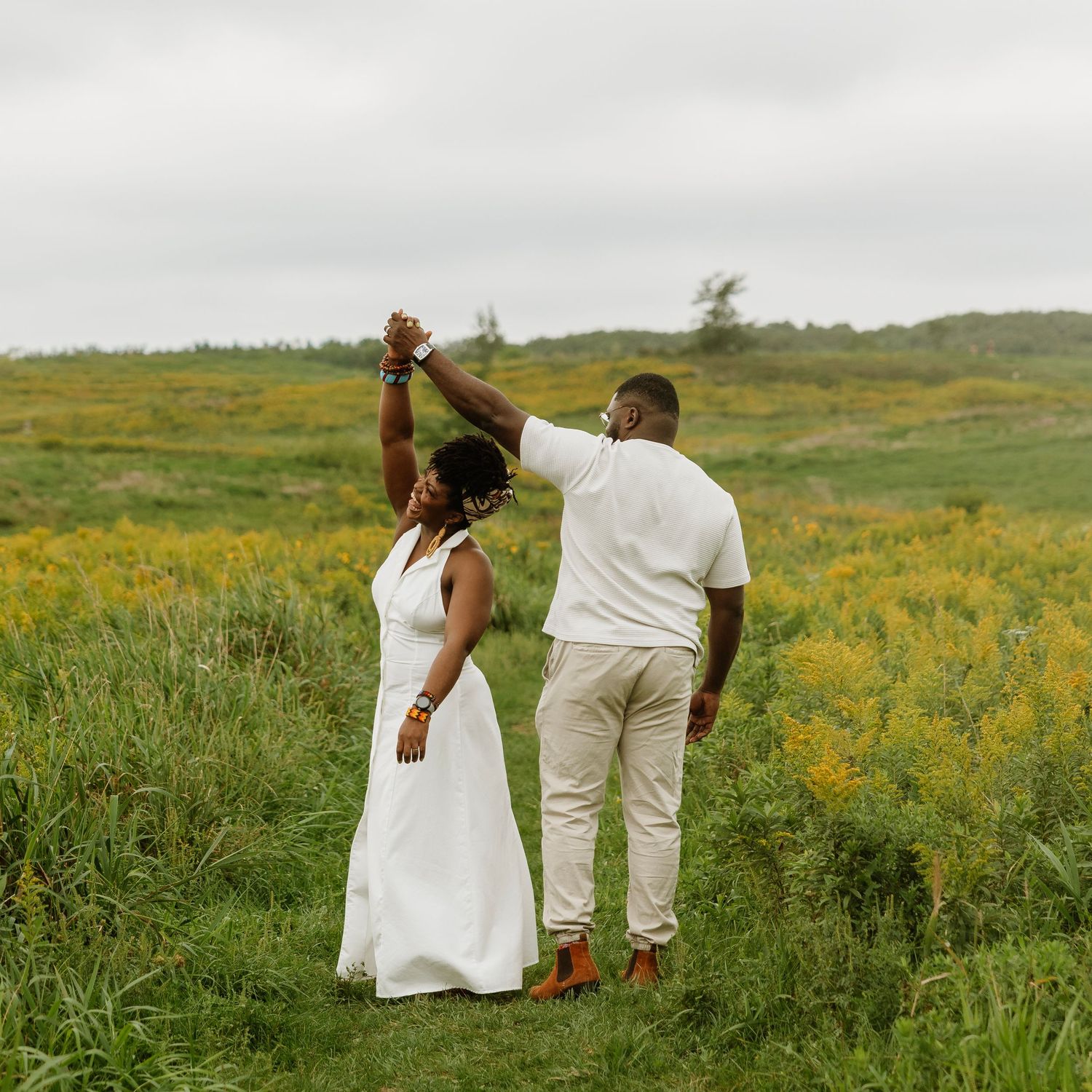 A couple dances in a lush green field, the woman in a white dress and man in white shirt and khaki pants.