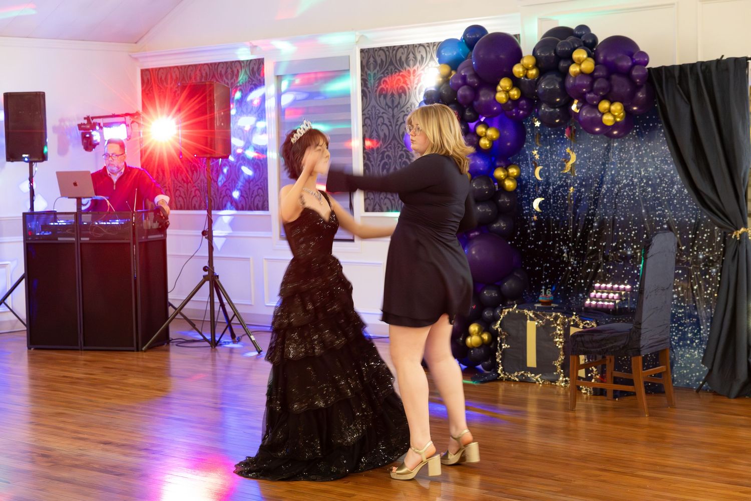 Two women dance at a prom event with a DJ, balloon arch, and celestial decorations in the background.