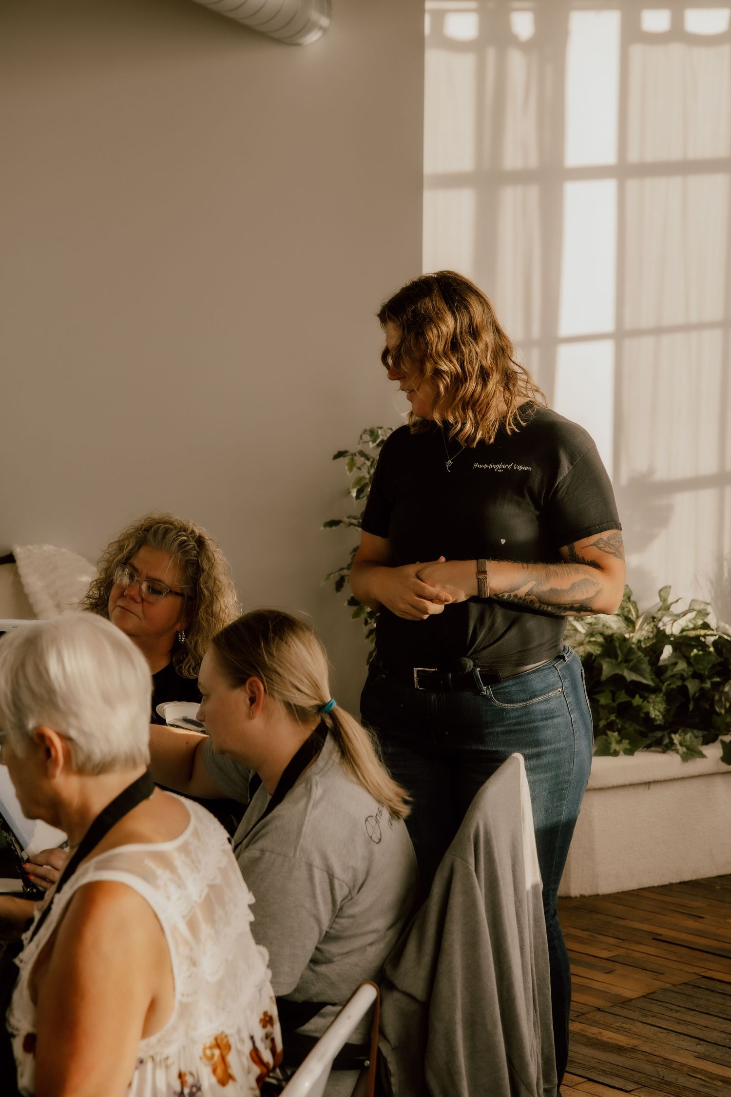 A standing woman in black shirt and jeans speaks to seated women in a bright, plant-filled indoor space.