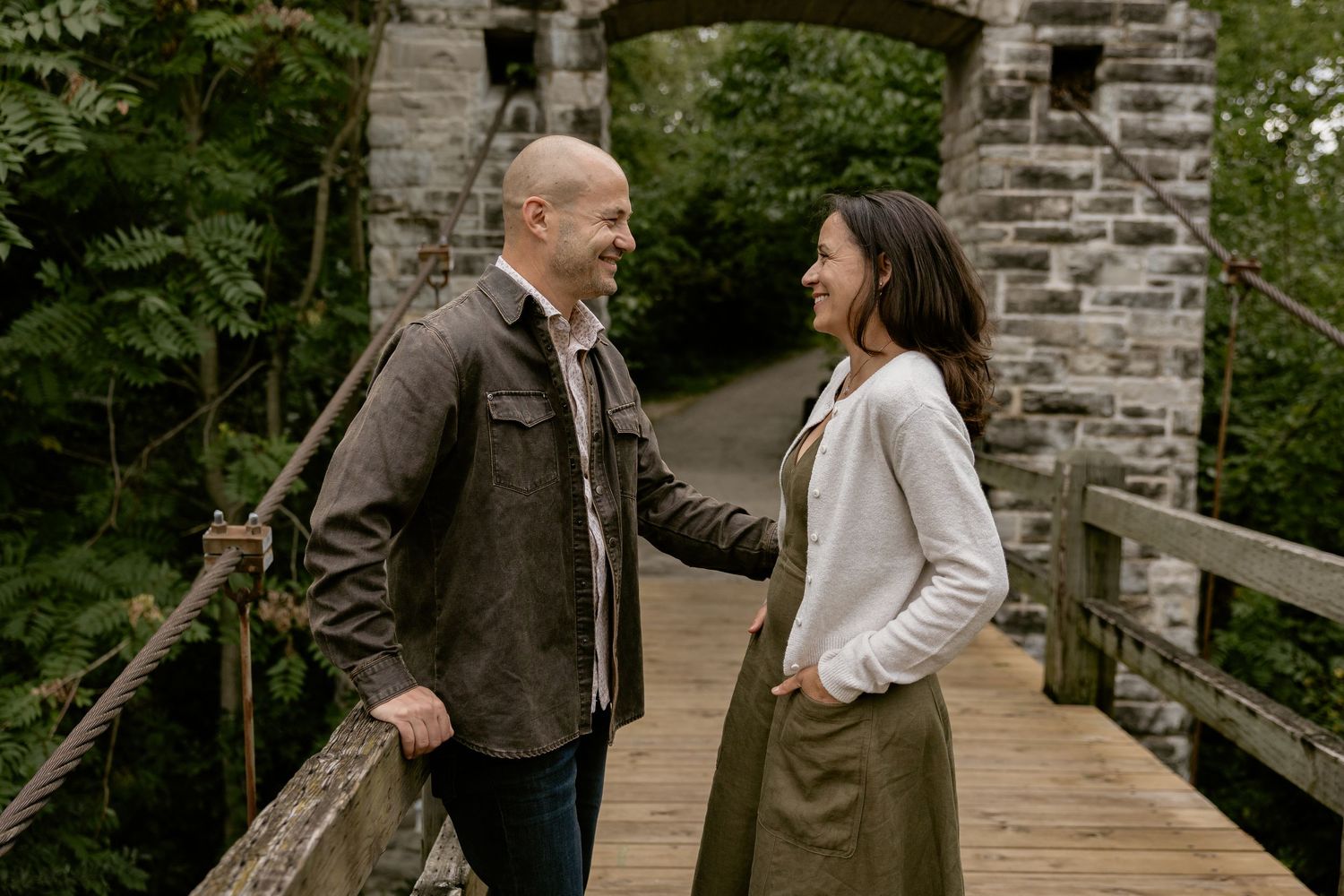 Couple smiling at each other on a wooden bridge surrounded by lush greenery and a stone archway.