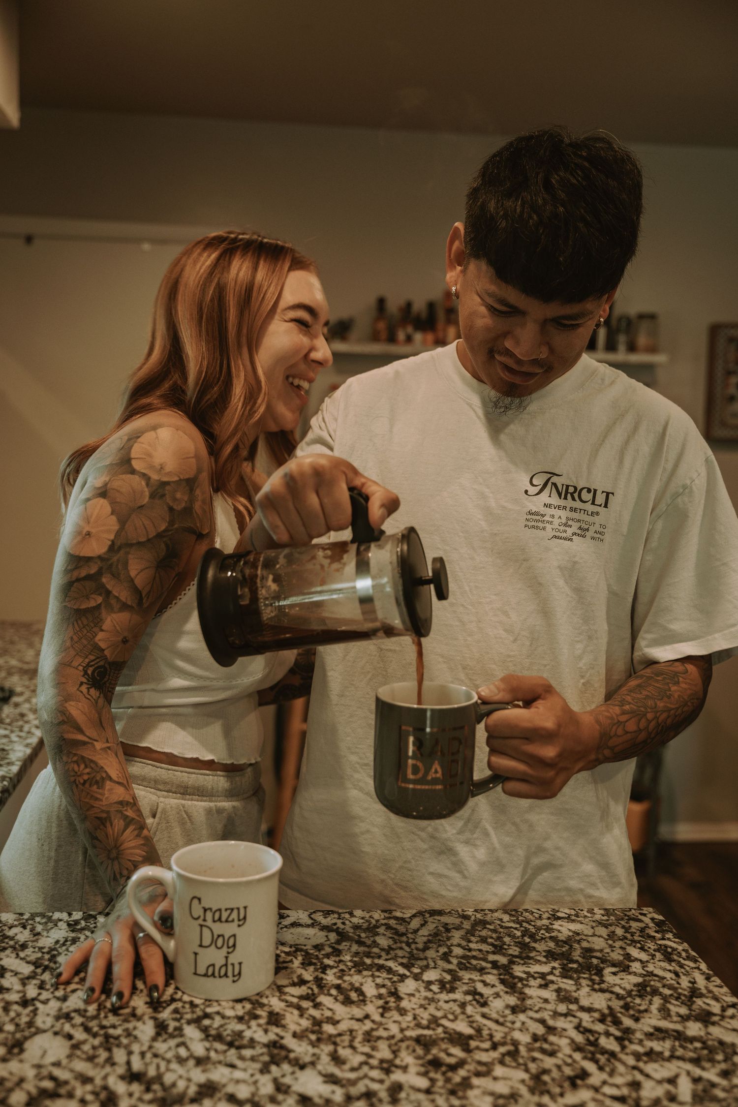 Couple laughing while pouring coffee from French press into mug, both with tattoos, Crazy Dog Lady mug on counter.