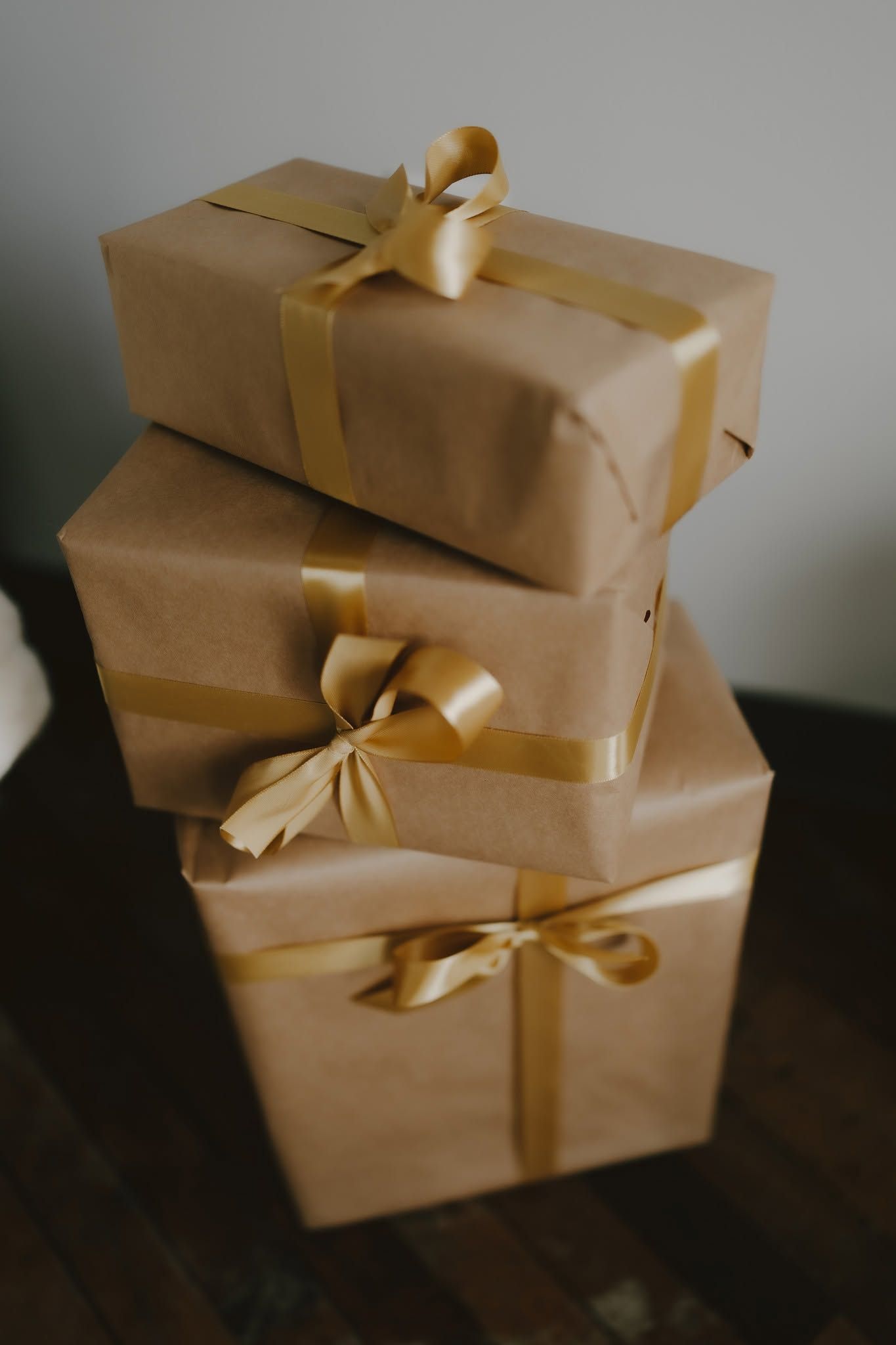 Three stacked kraft paper gift boxes tied with gold satin ribbons and bows on a dark wooden surface.