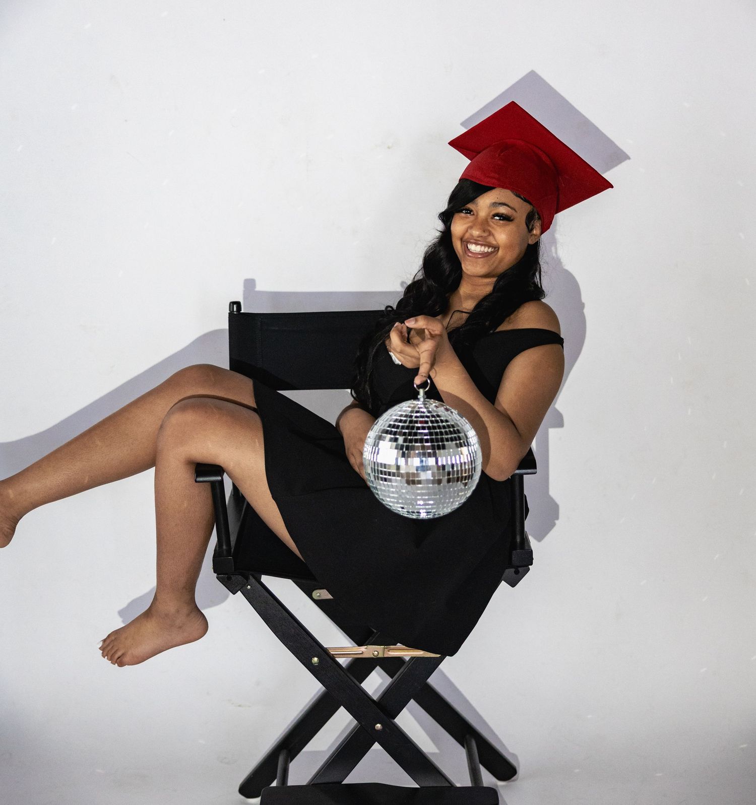 Young woman in graduation cap and black dress holding disco ball, sitting in director's chair, smiling.