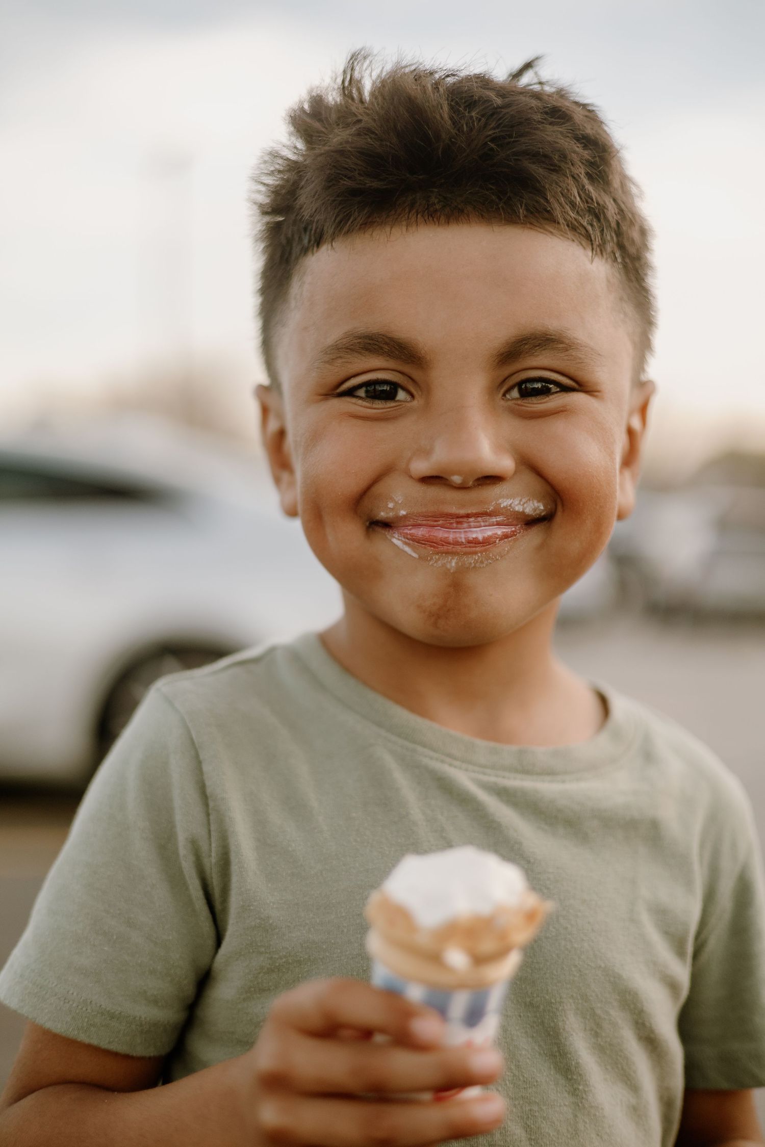 Smiling young boy with ice cream on his face holding a vanilla ice cream cone outdoors.