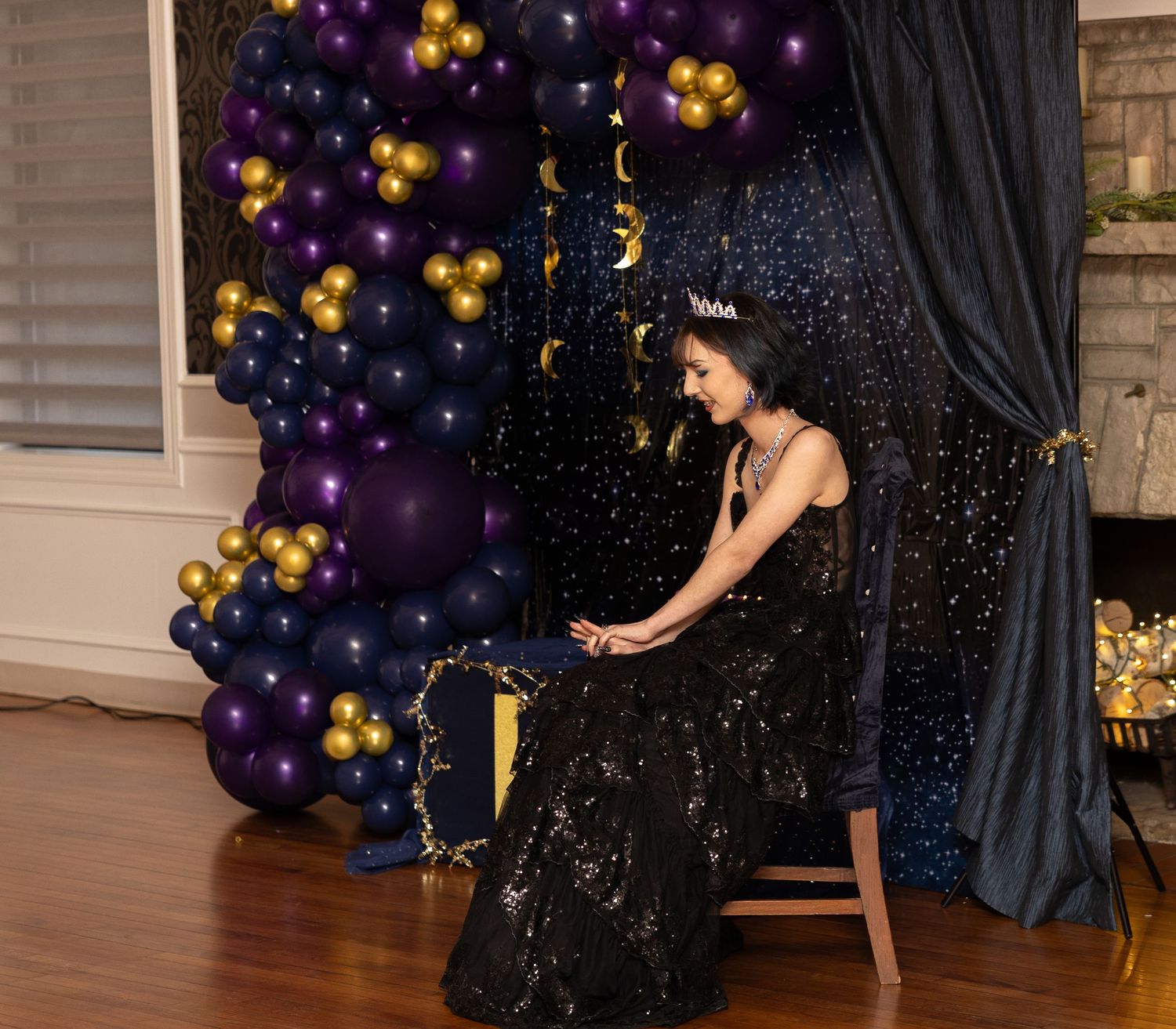 Young woman in black sequin gown wearing tiara seated before purple gold navy balloon arch backdrop.