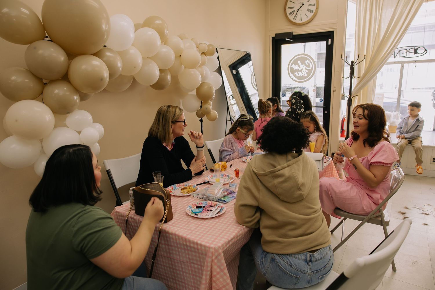 Women gathered around a pink gingham table for a craft party, decorated with white balloon garland indoors.