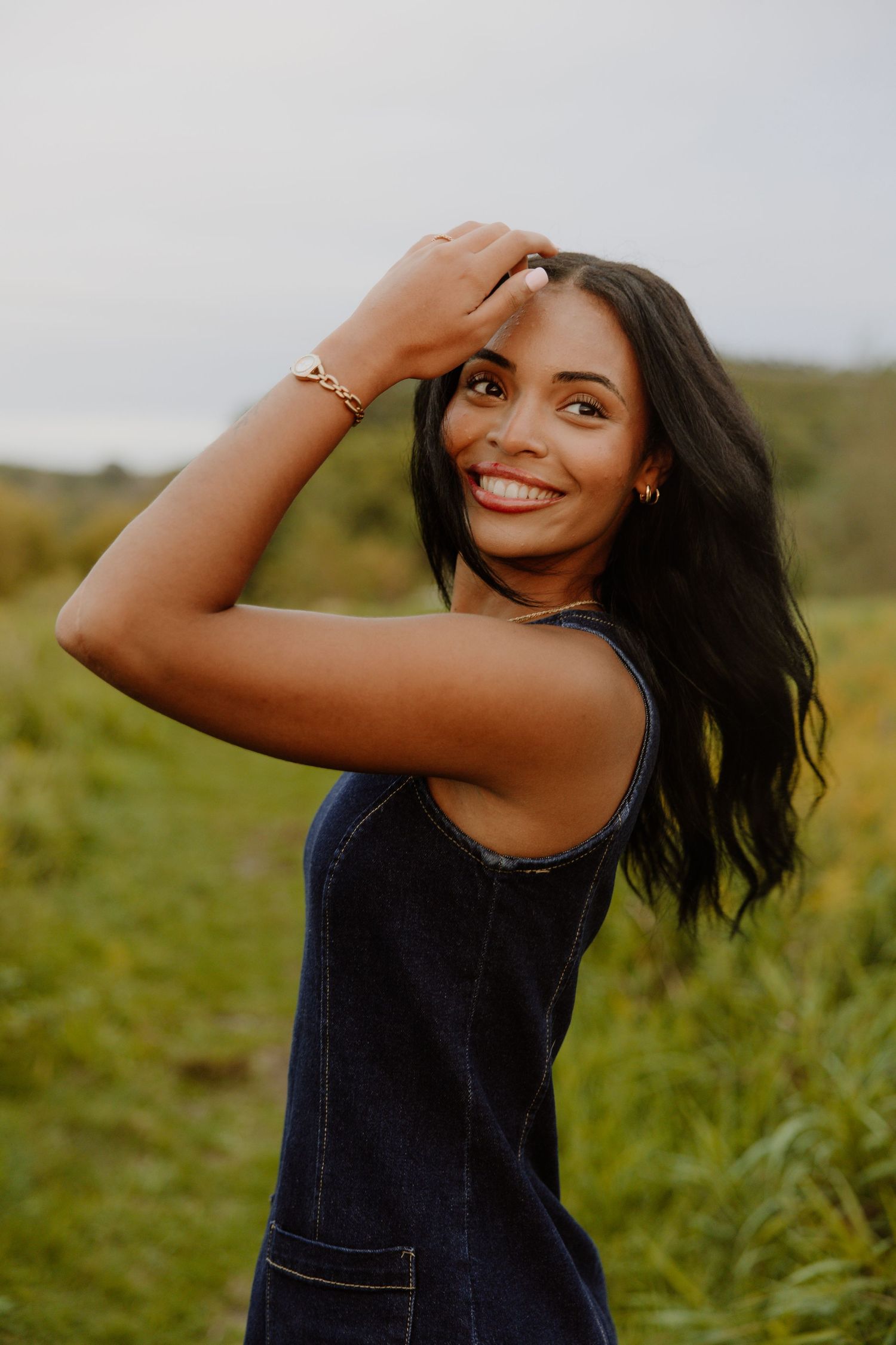 Smiling young woman in denim dress posing outdoors in green field, wearing gold bracelet with hand raised to head.