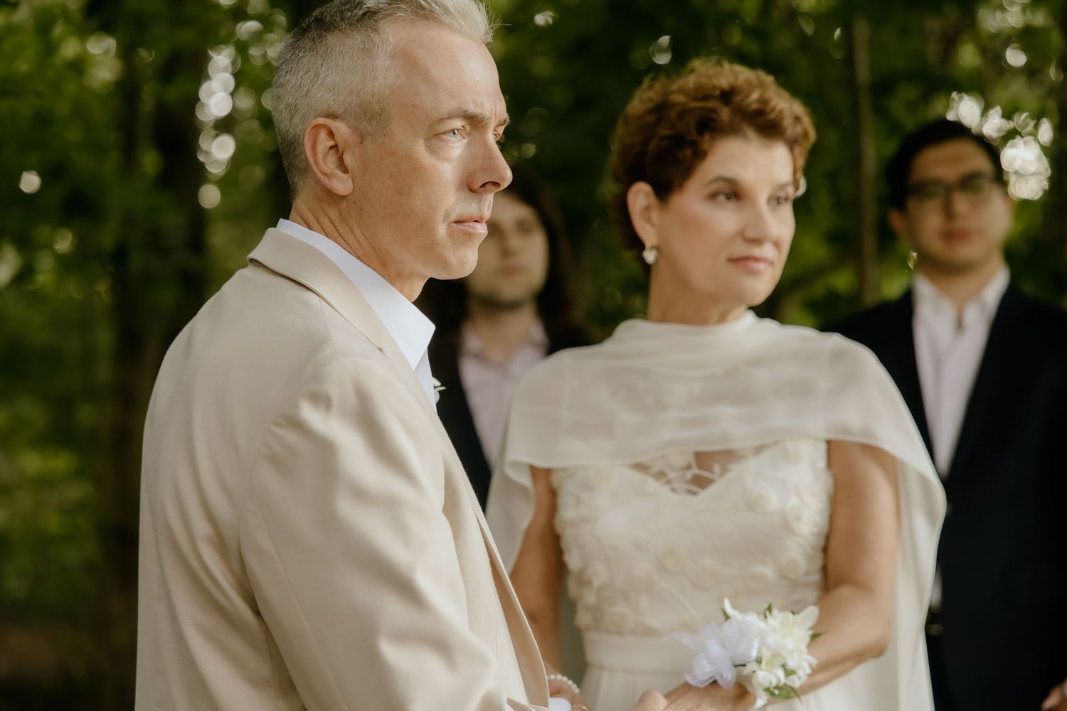 Elegant outdoor wedding ceremony with bride in white cape dress and groom in cream suit standing among lush green trees.