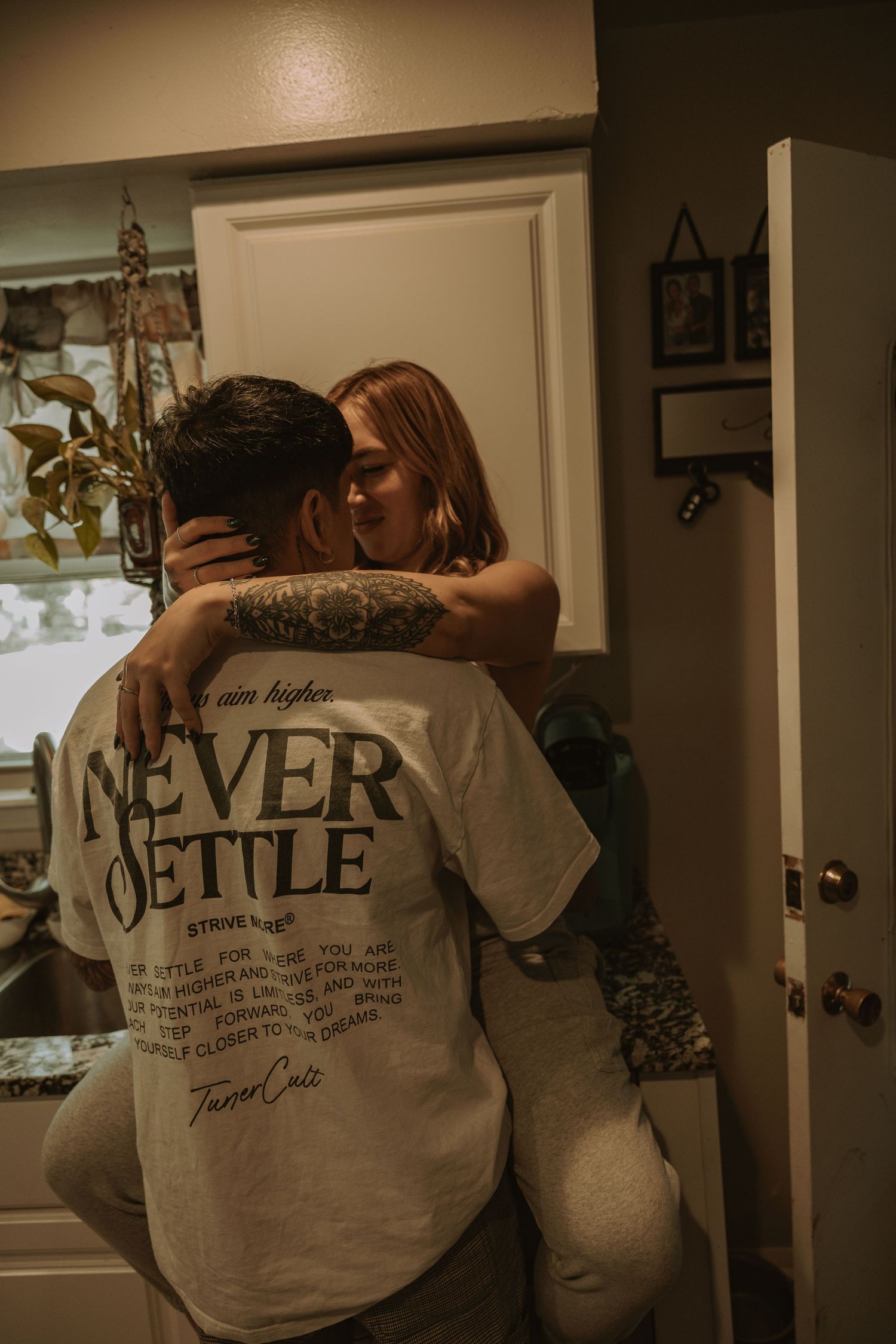 Couple embracing in kitchen, man wearing Never Settle graphic tee with tattoos, woman with red hair hugging him tenderly.