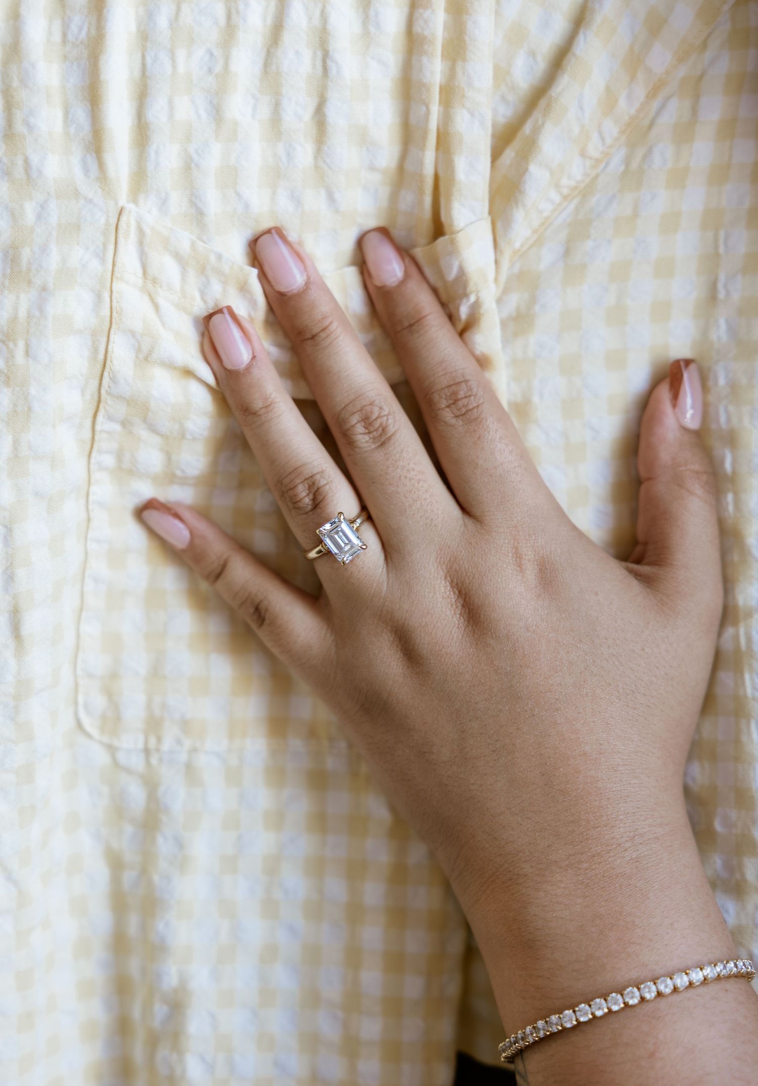 Hand wearing an emerald-cut diamond solitaire engagement ring with a tennis bracelet, resting on a yellow gingham shirt.