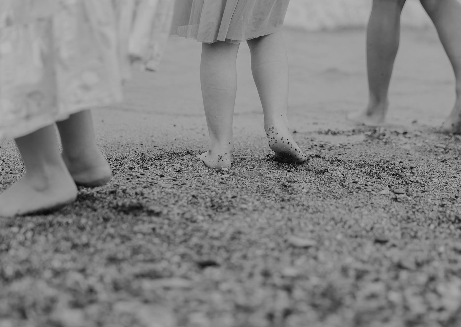 Black and white photo of barefoot children standing on sandy beach, wearing dresses, captured from the knees down.