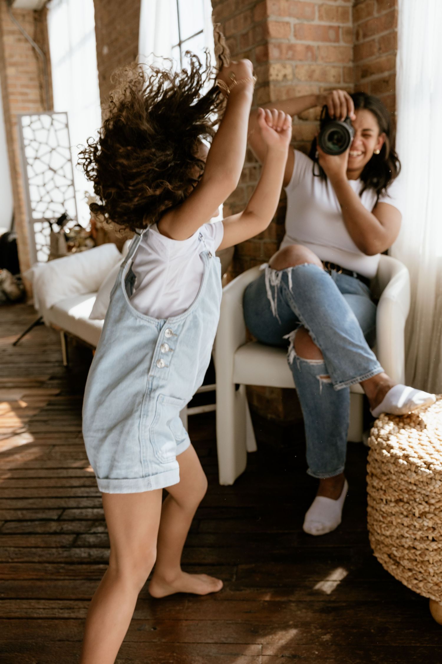 A smiling woman photographs a curly-haired child dancing in denim overalls inside a cozy brick-walled room.