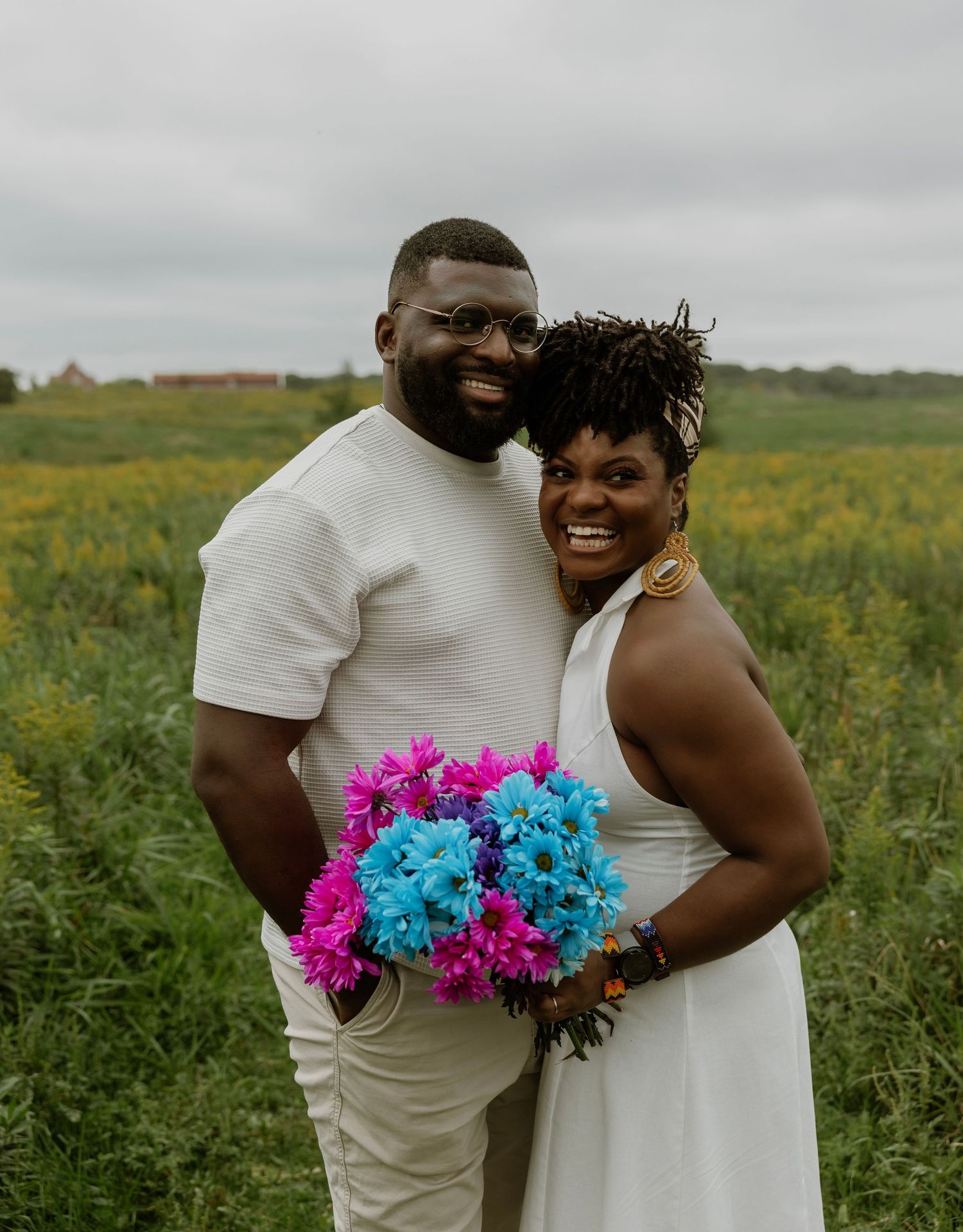 Couple in white attire smiling outdoors in a green field, woman holding vibrant pink and blue flower bouquet.