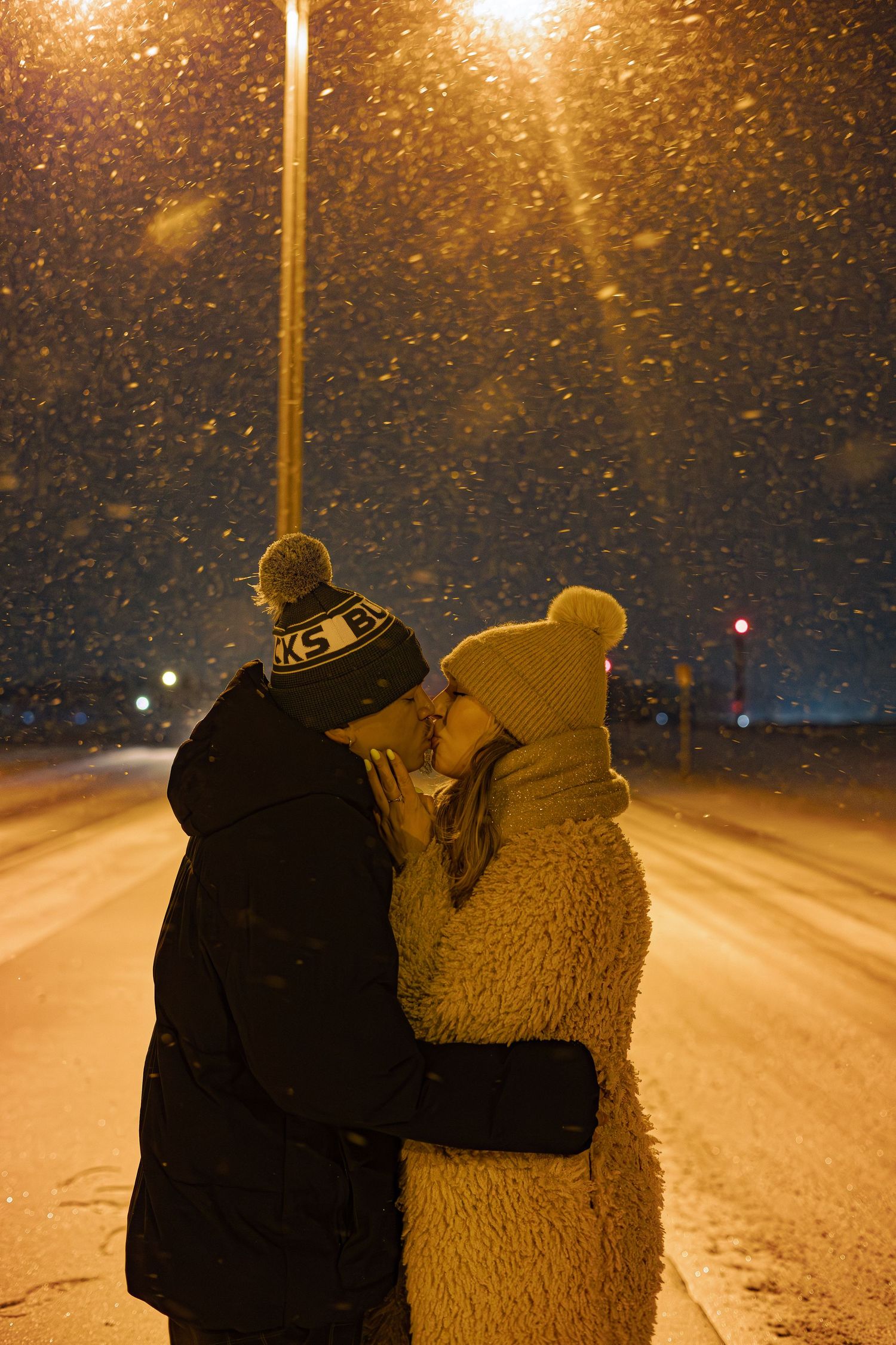 Couple kissing in snowfall at night under street lights, wearing winter hats and coats on a snowy road.