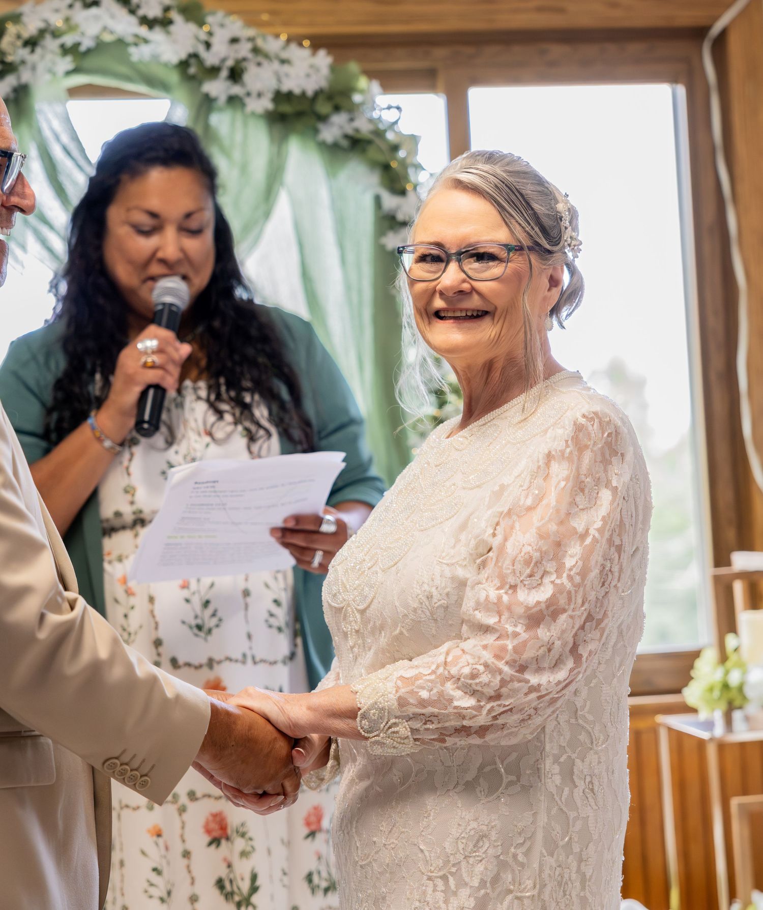 Smiling bride in white lace dress holds hands at wedding ceremony while officiant reads vows indoors.