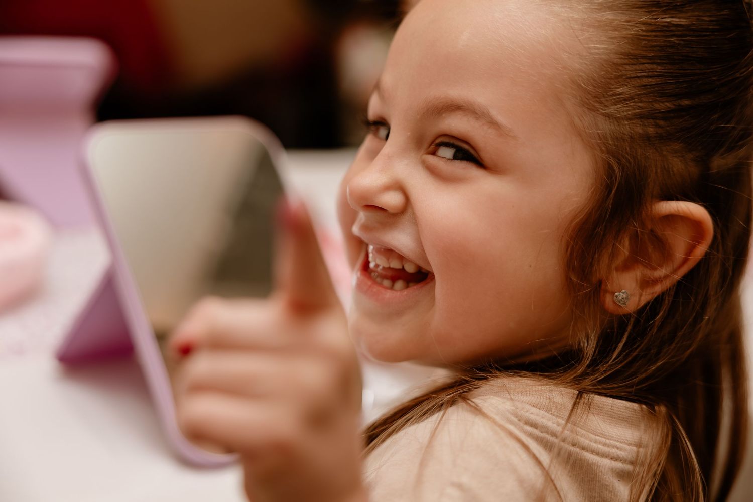 A happy young girl smiles and gives a thumbs up while using a pink tablet device.