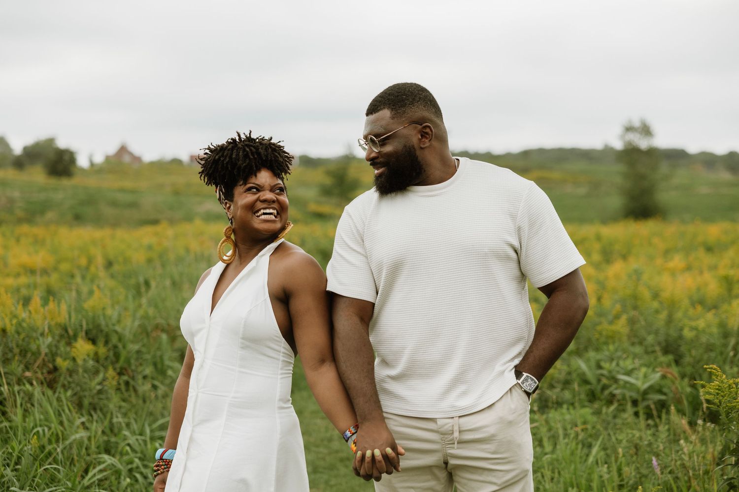 A joyful Black couple holds hands and smiles in a lush green field with yellow wildflowers on a cloudy day.
