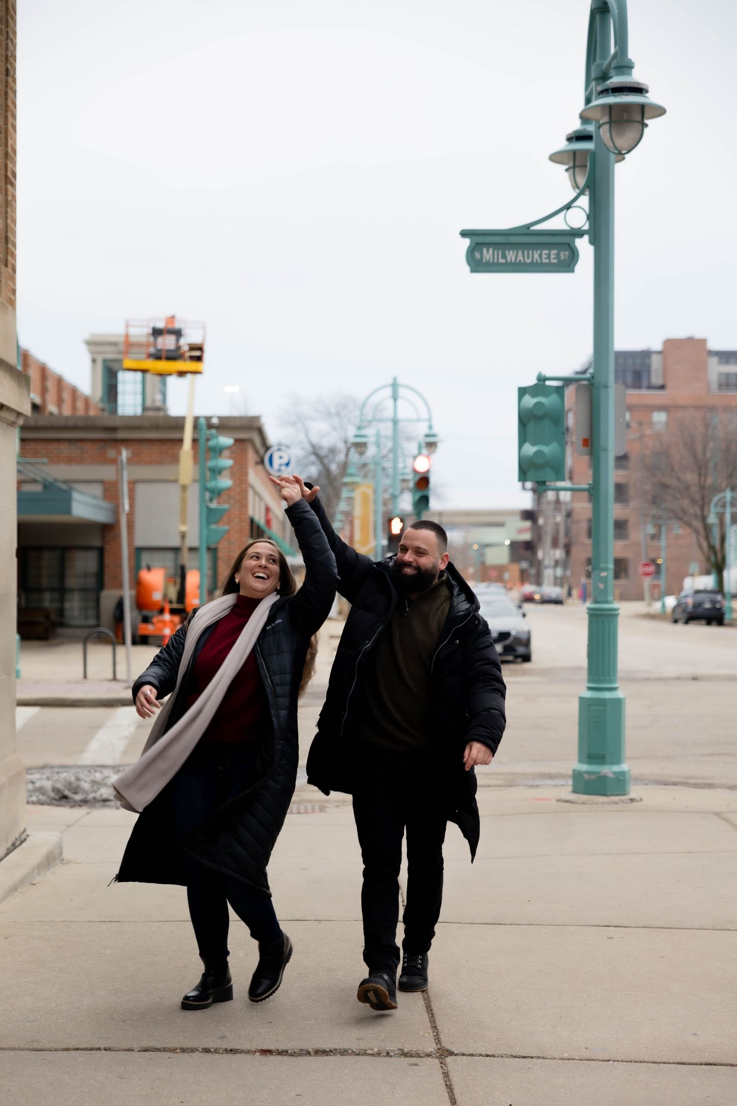 Couple dancing joyfully on a Milwaukee street corner in winter coats on a cloudy day.