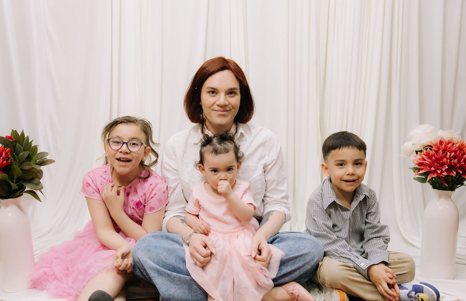 A woman sits with three young children in front of a white curtain backdrop with pink floral arrangements.