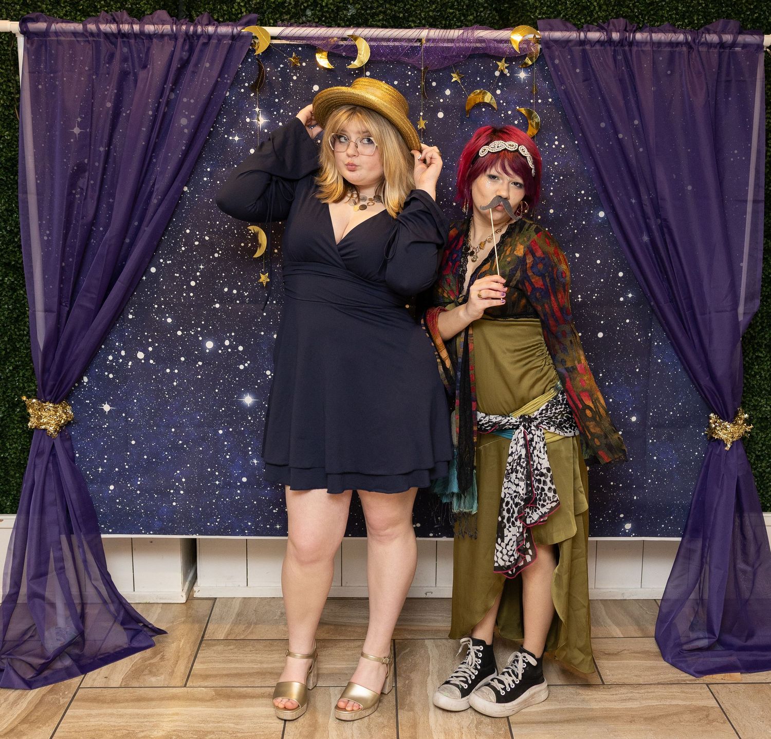 Two women in costumes pose in front of a starry backdrop with purple curtains at a themed photo booth.
