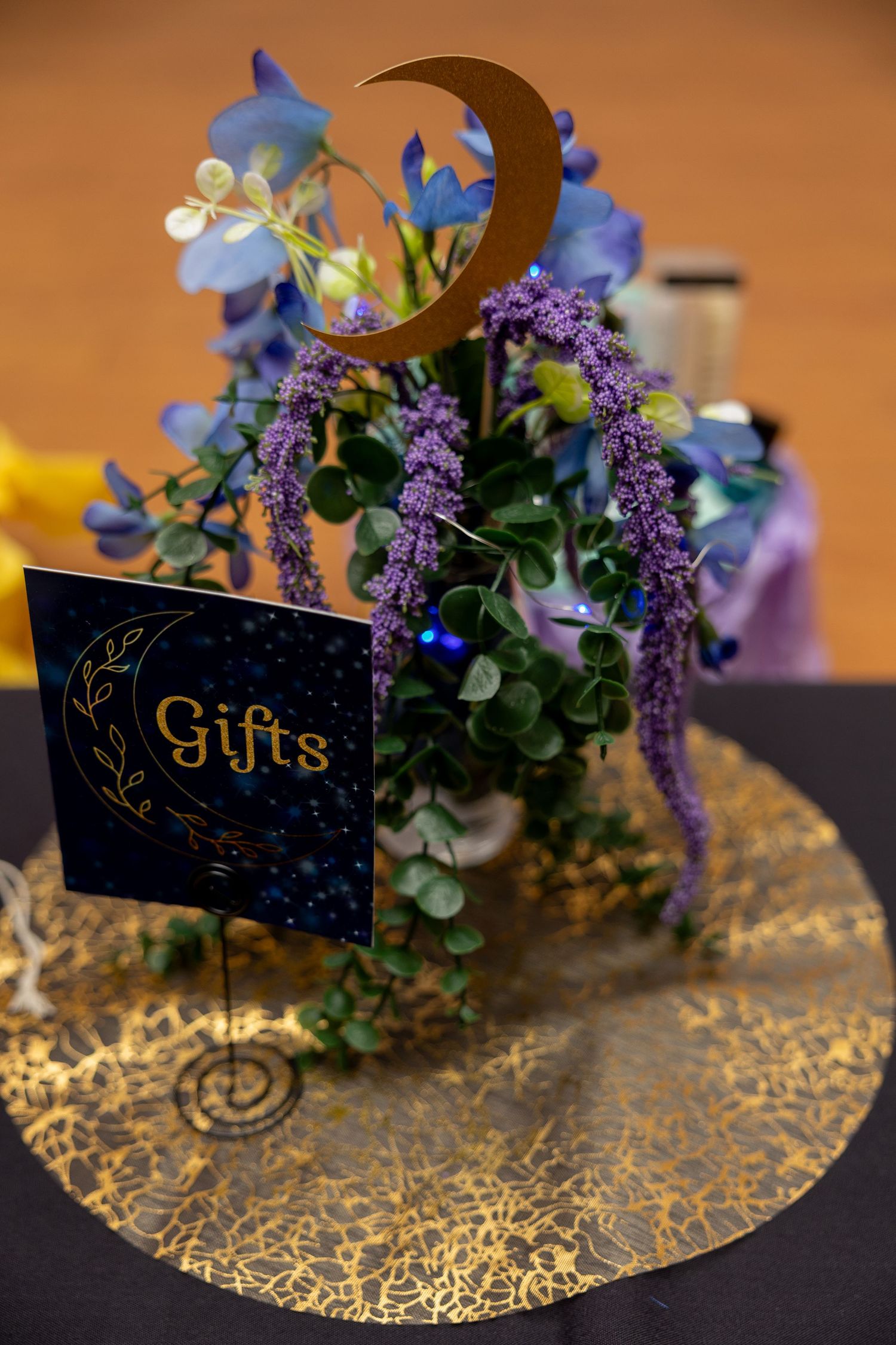 Gold crescent moon wedding gifts table sign with purple cascading flowers and blue blooms on gold decorative plate.