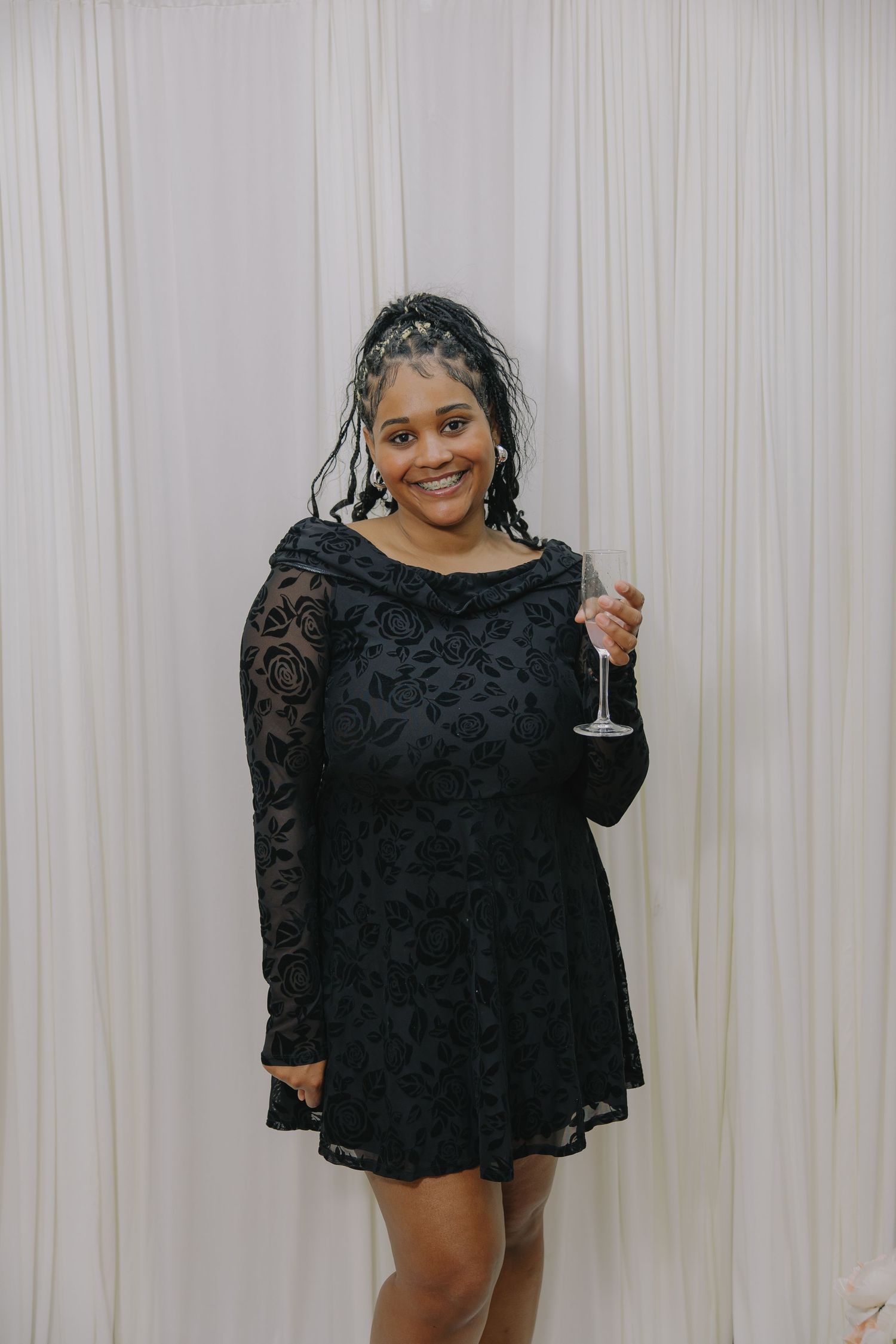 Woman in black lace dress holding champagne glass, smiling against white drape backdrop.