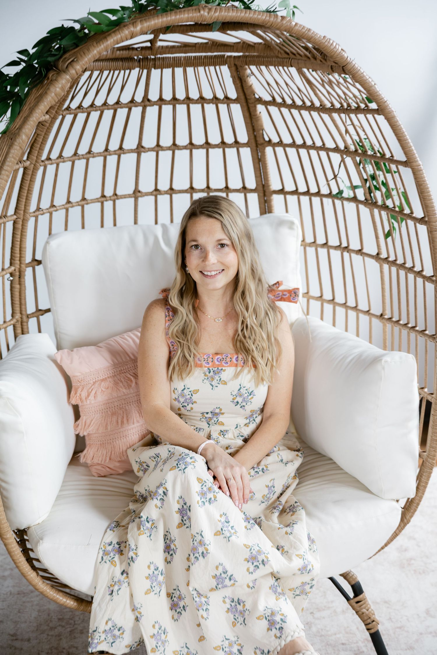 Young woman in floral dress sitting in a rattan egg chair with white cushions and pink pillow.