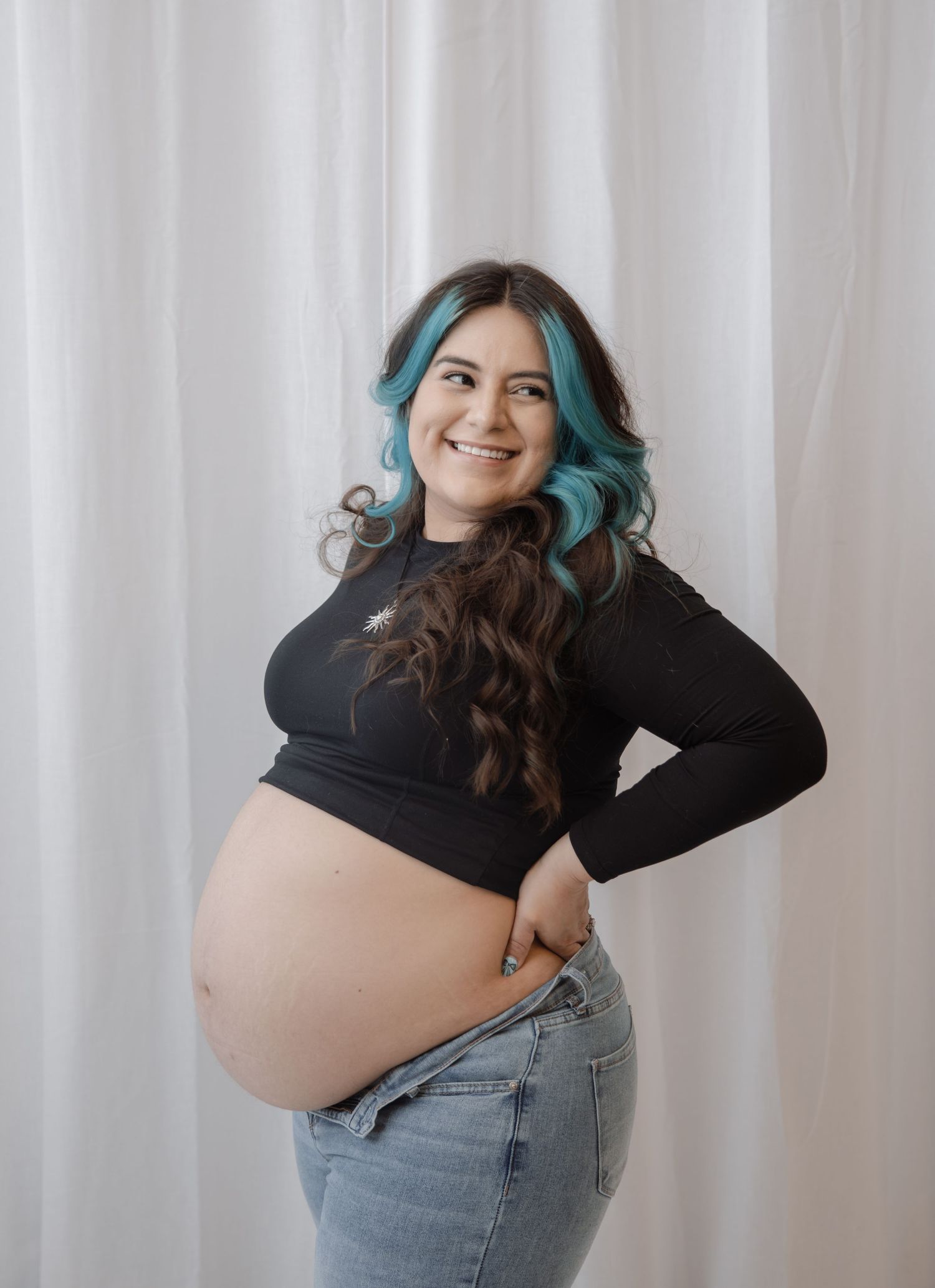 Smiling pregnant woman with blue-tipped hair in black crop top and jeans posing against white curtain backdrop.