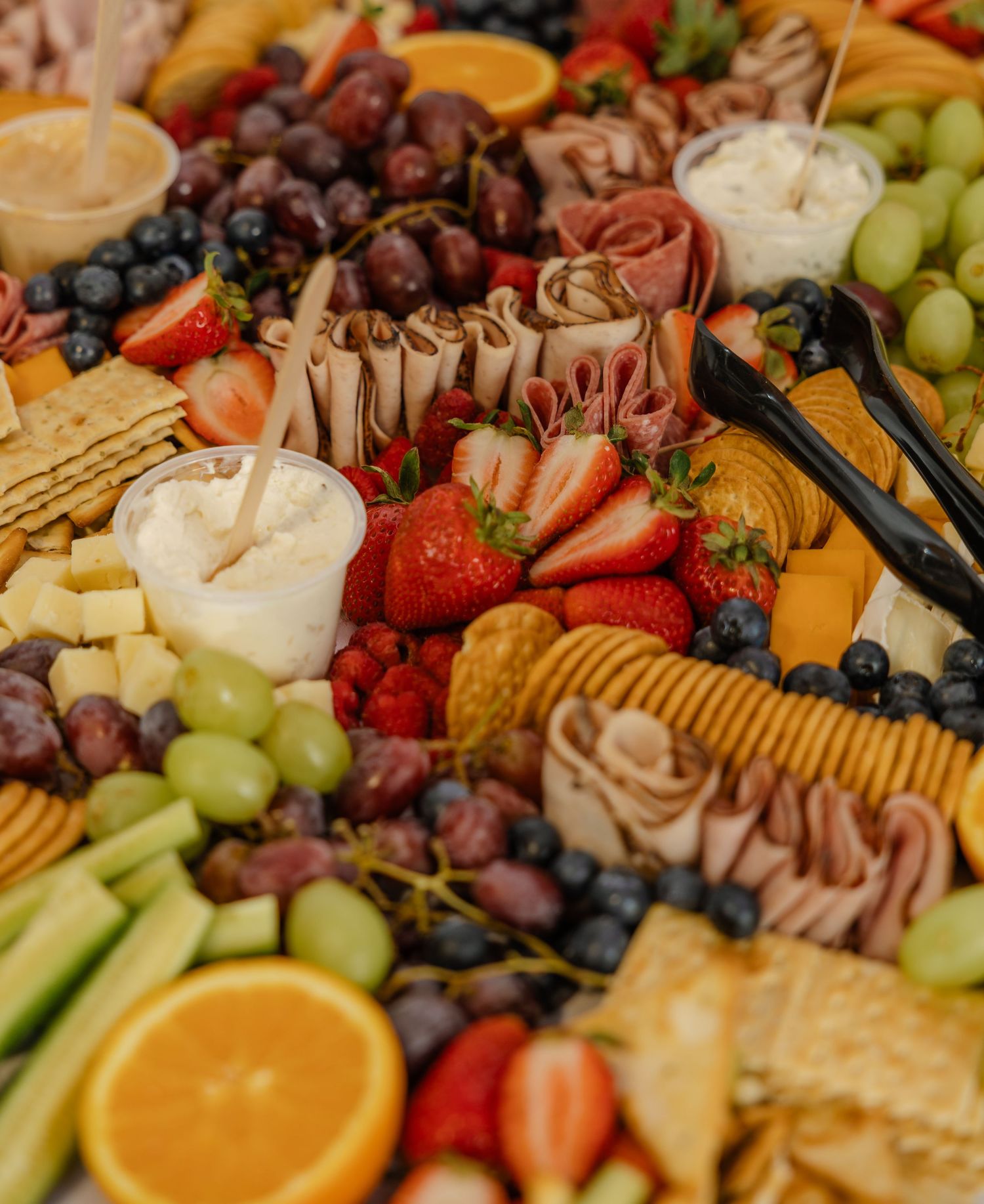Colorful charcuterie board with strawberries, grapes, blueberries, oranges, deli meats, cheeses, crackers, and dipping sauces.