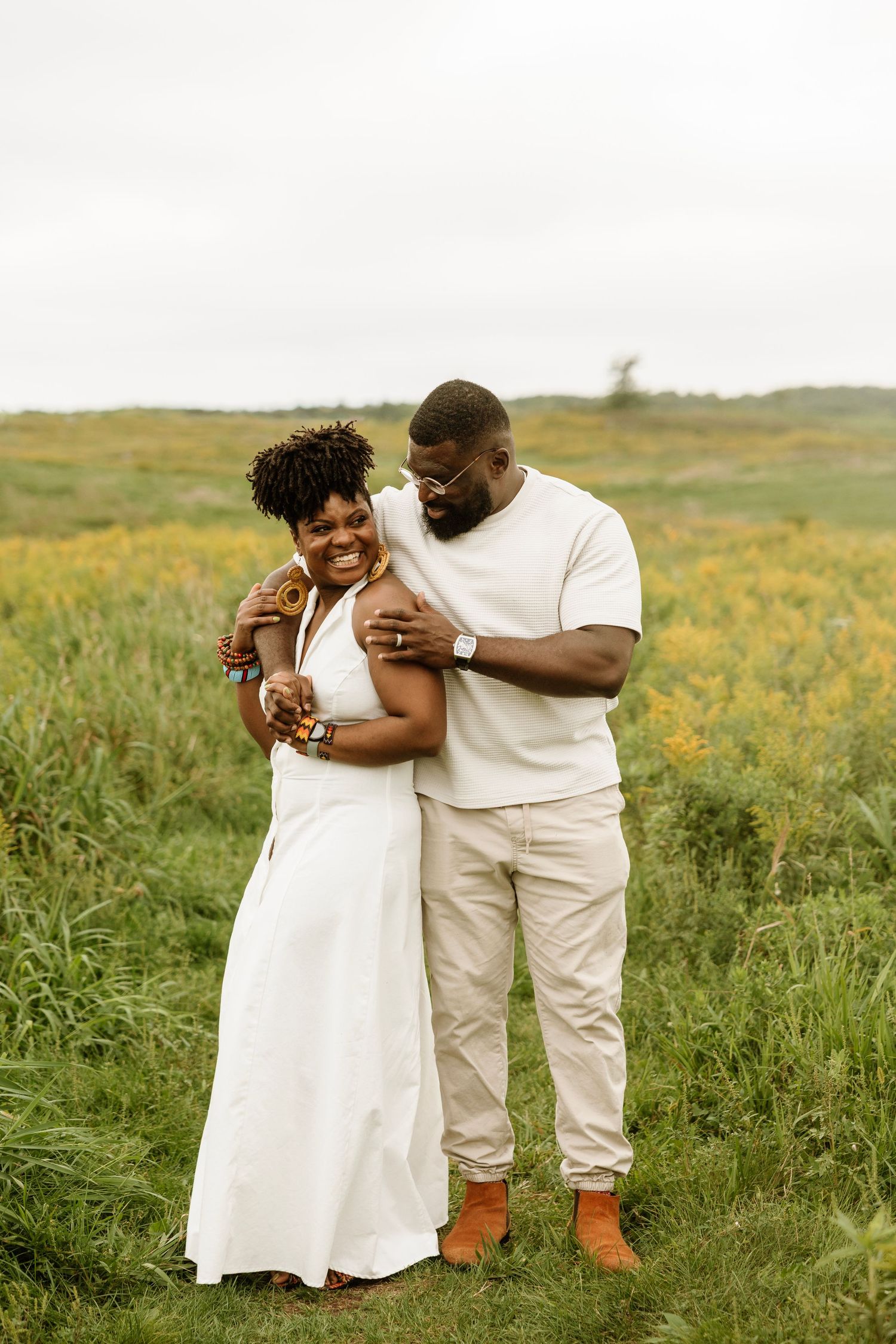 A joyful Black couple embraces in a lush green field with yellow wildflowers, dressed in coordinating white and cream outfits.