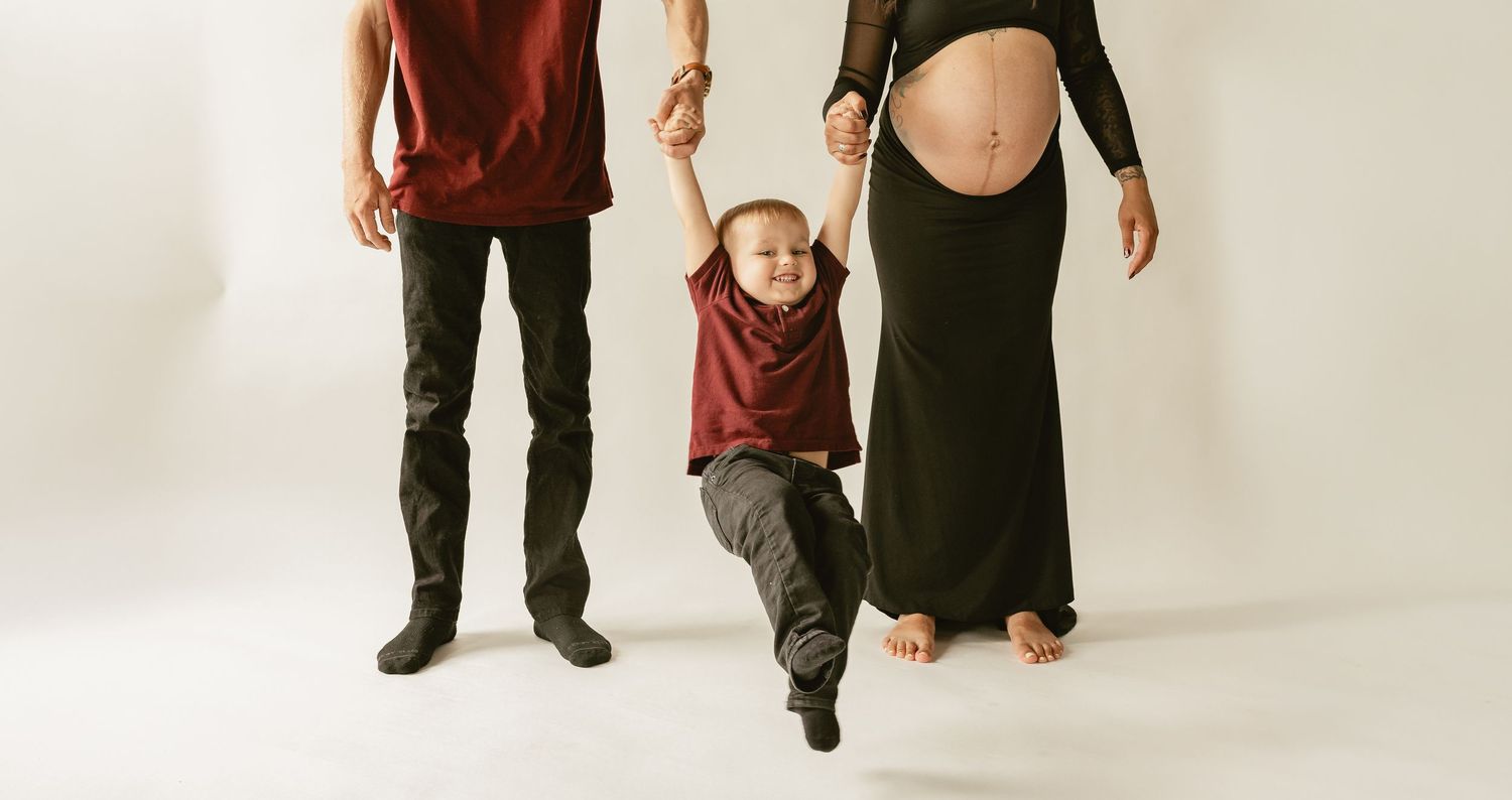 Parents hold hands of toddler boy swinging between them, mother visibly pregnant in black dress, white studio background.
