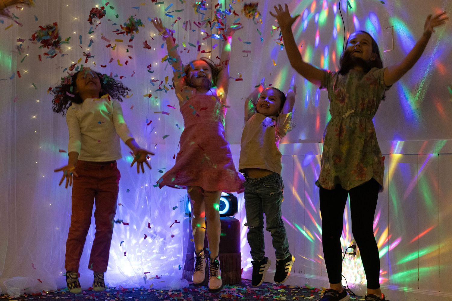 Four children joyfully tossing confetti under colorful disco lights at a festive party celebration.