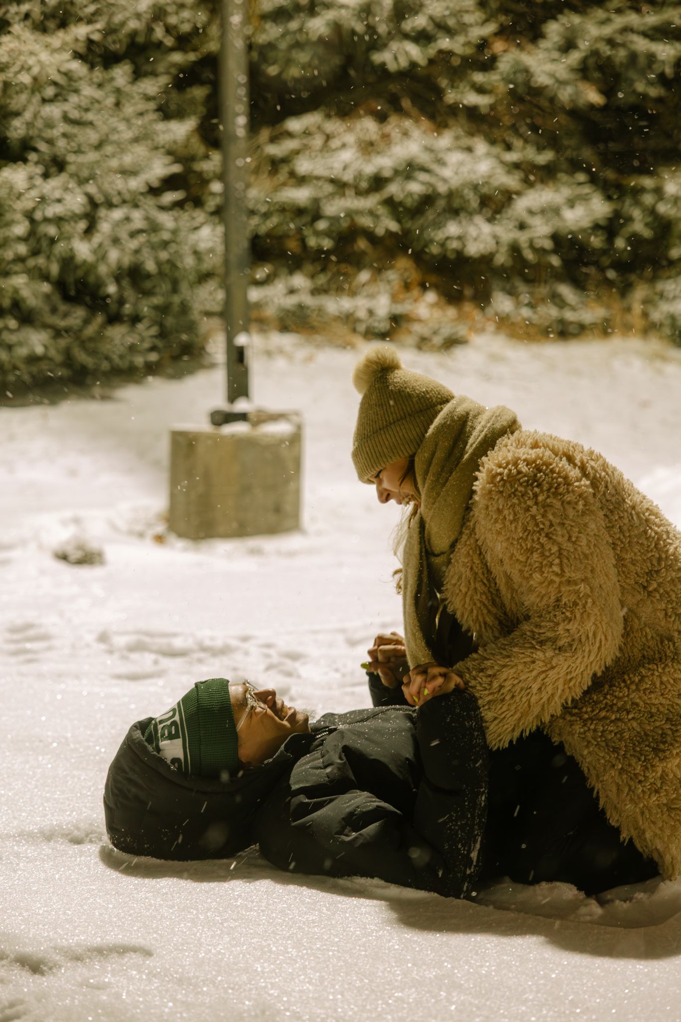 Two people playing in the snow, one lying down while the other kneels beside them, both wearing winter coats and hats.