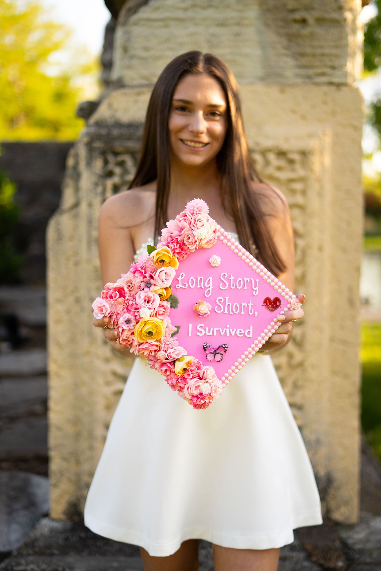 Smiling graduate in white dress holds pink floral decorated cap reading Long Story Short I Survived outdoors.