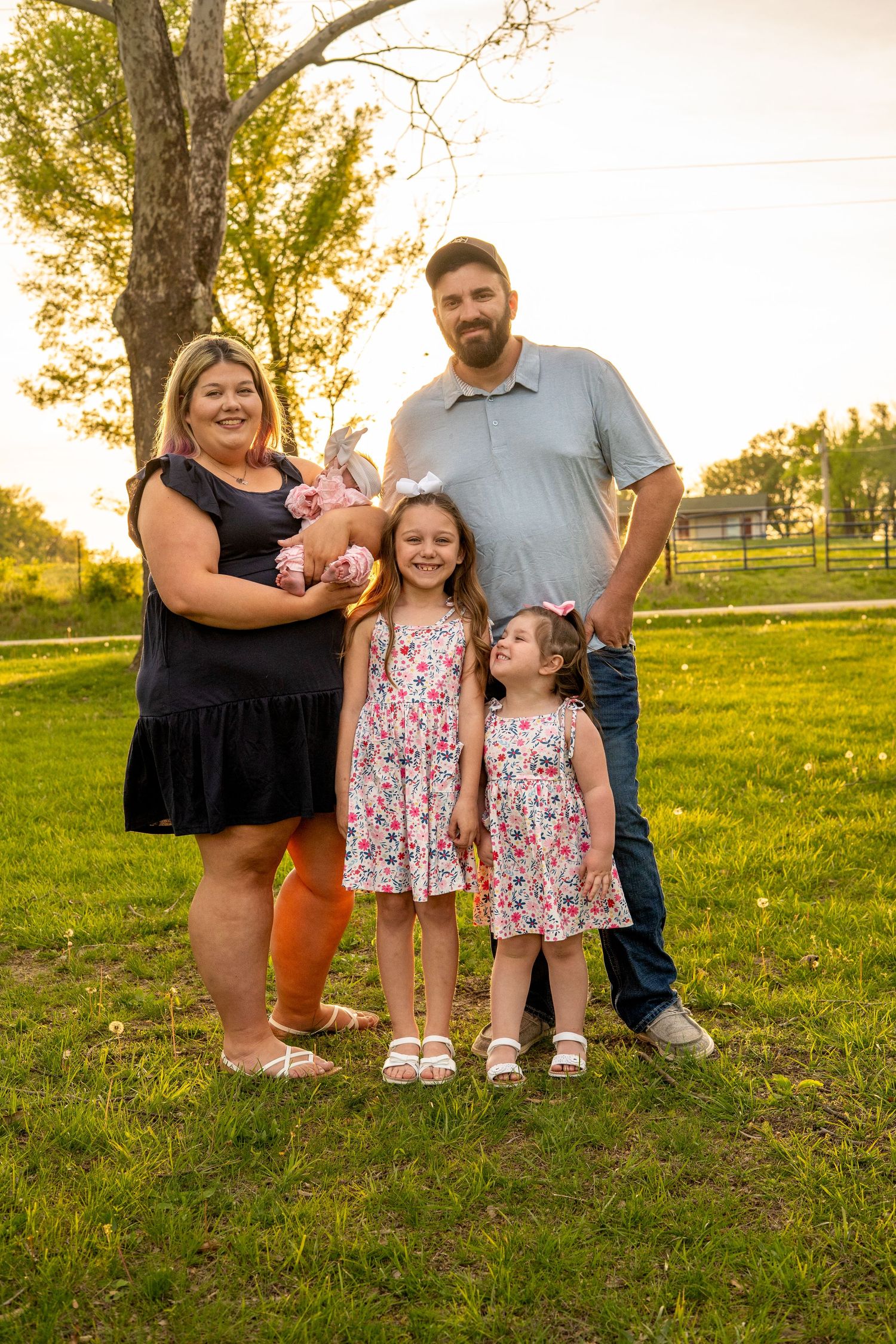 Happy family of five with two young girls in floral dresses and a newborn posing in a green grassy field.