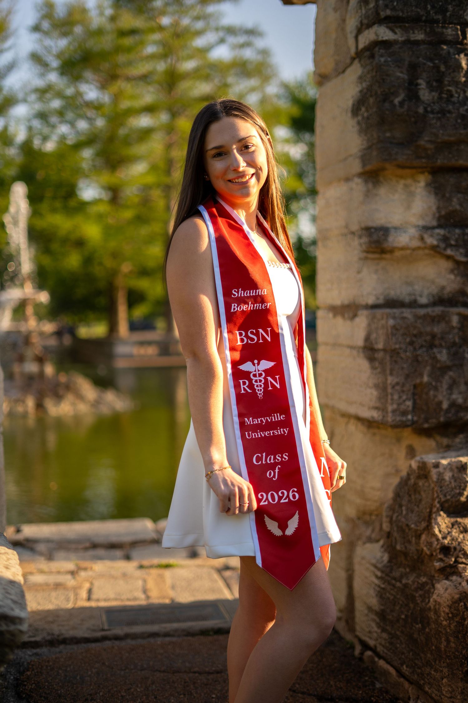 Nursing graduate in white dress wears red BSN RN Maryville University Class of 2026 stole near stone column.