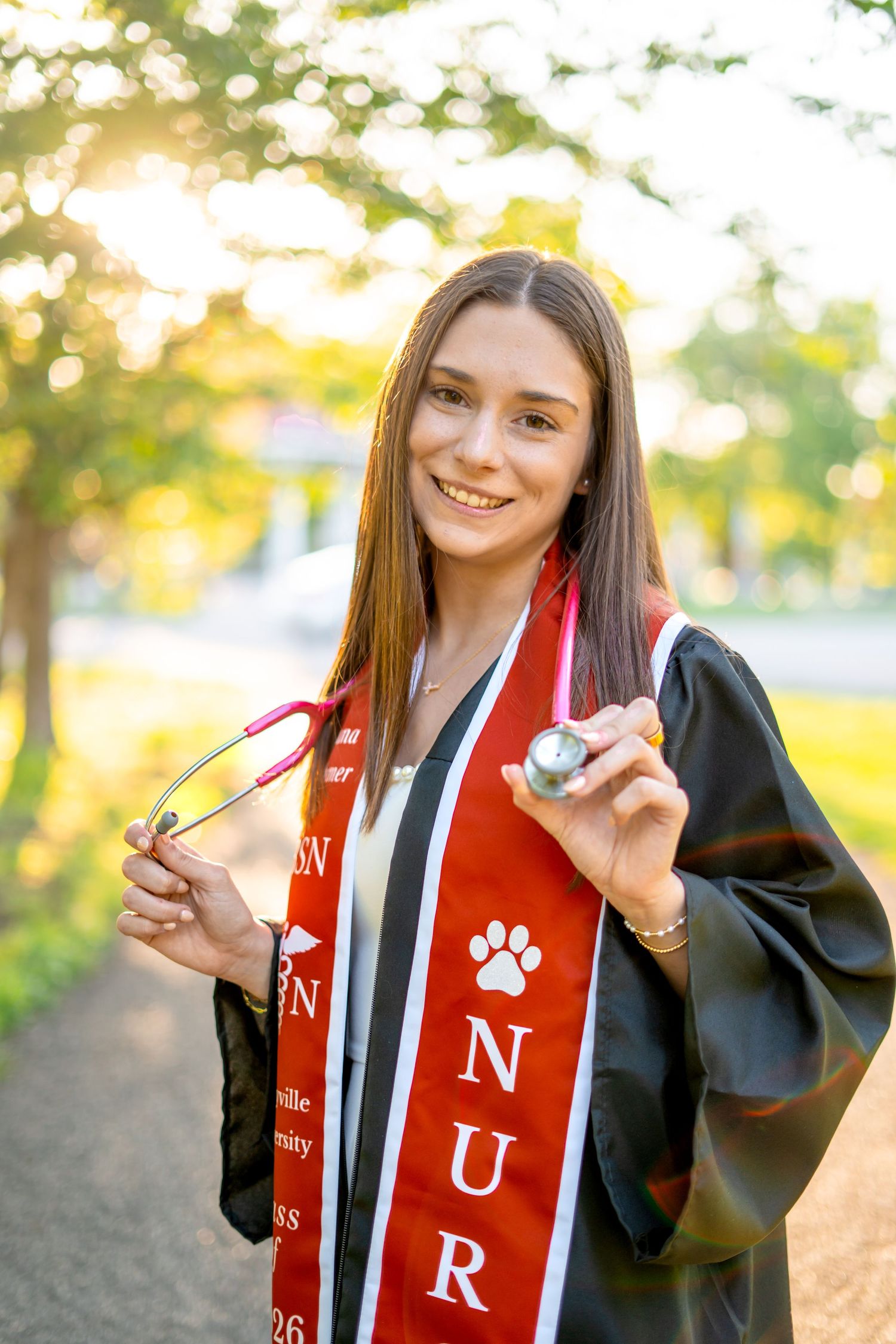 Smiling nursing graduate holds stethoscope wearing black gown and red nursing honor stole.