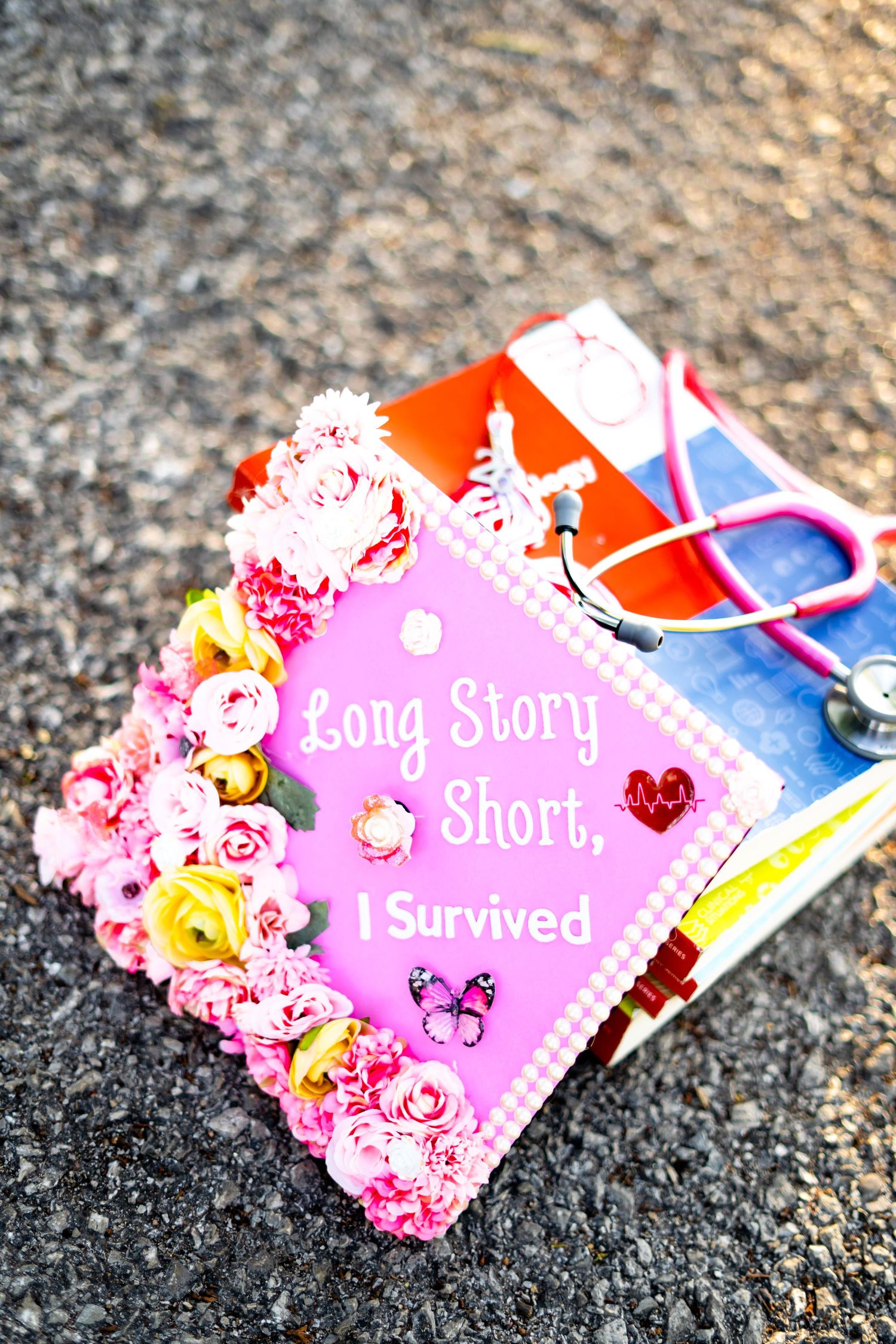 Decorated pink graduation cap reading Long Story Short I Survived with flowers and a stethoscope on pavement.