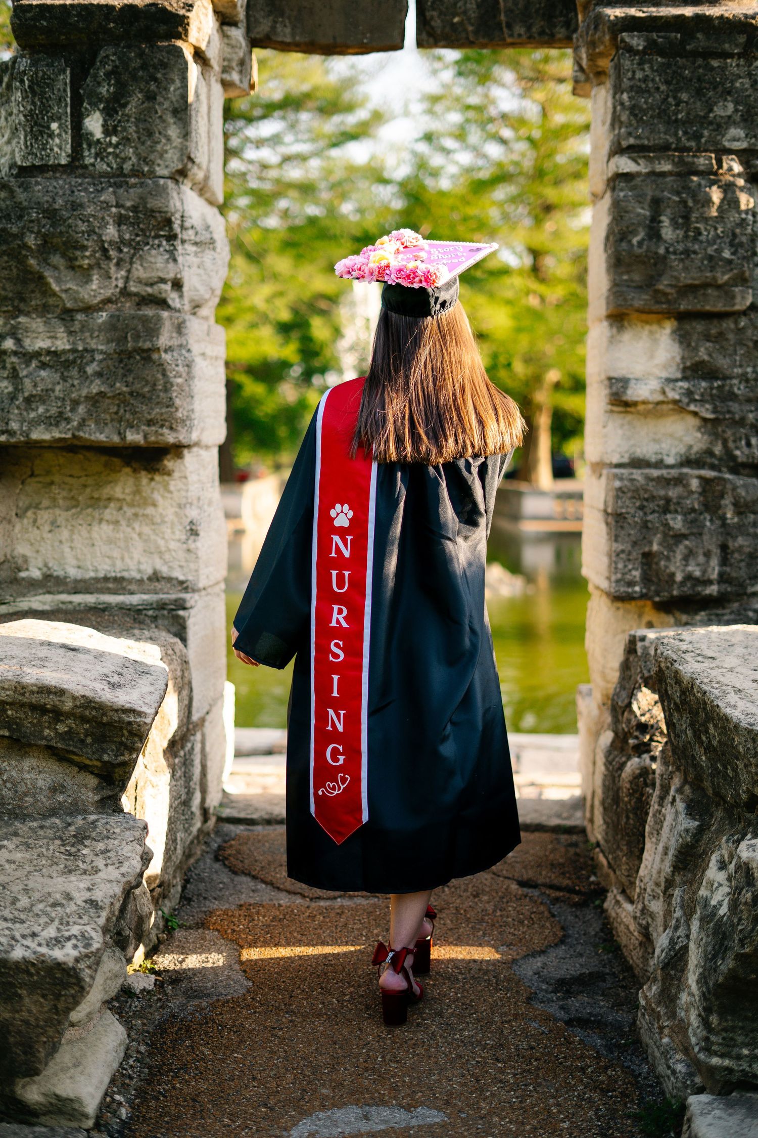 Nursing graduate in black gown with red nursing sash walking through stone arch toward fountain.